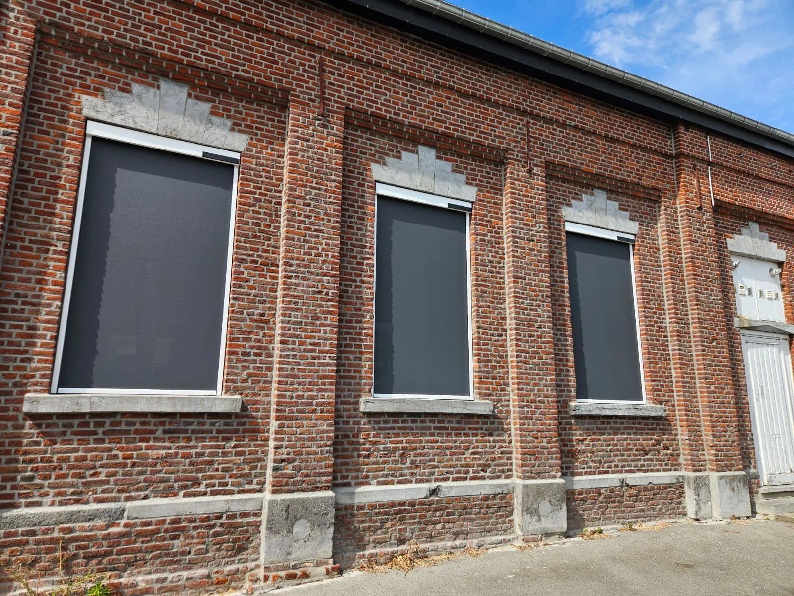 Façade d'un bâtiment en briques rouges avec trois fenêtres à volets roulants gris foncé et encadrements en pierre, sous un ciel bleu avec quelques nuages.