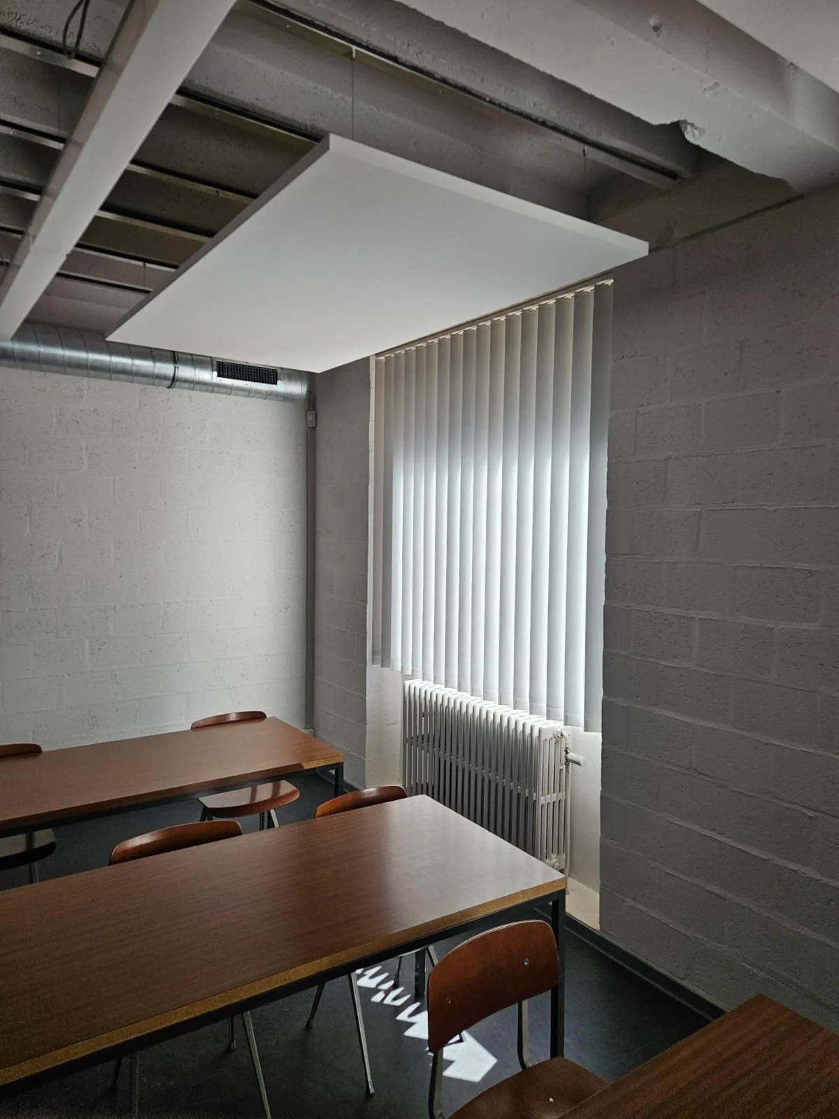Une salle de réunion ou de classe avec des tables en bois et des chaises, un mur en briques peintes en blanc, une grande fenêtre avec des voilages verticaux et un radiateur.