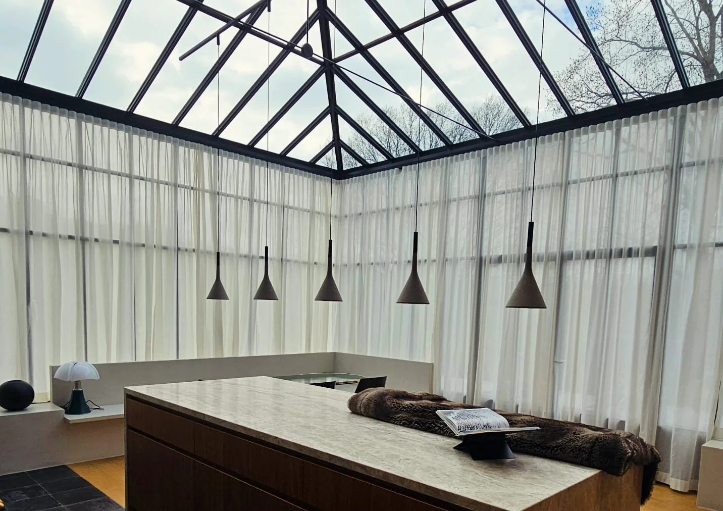 Intérieur d'une pièce avec de grandes fenêtres vitrées couvertes de rideaux blancs, un plafond en verre avec une structure en métal, un comptoir en bois avec une surface en marbre, un tapis en fourrure, une lampe de table et une suspension noire.