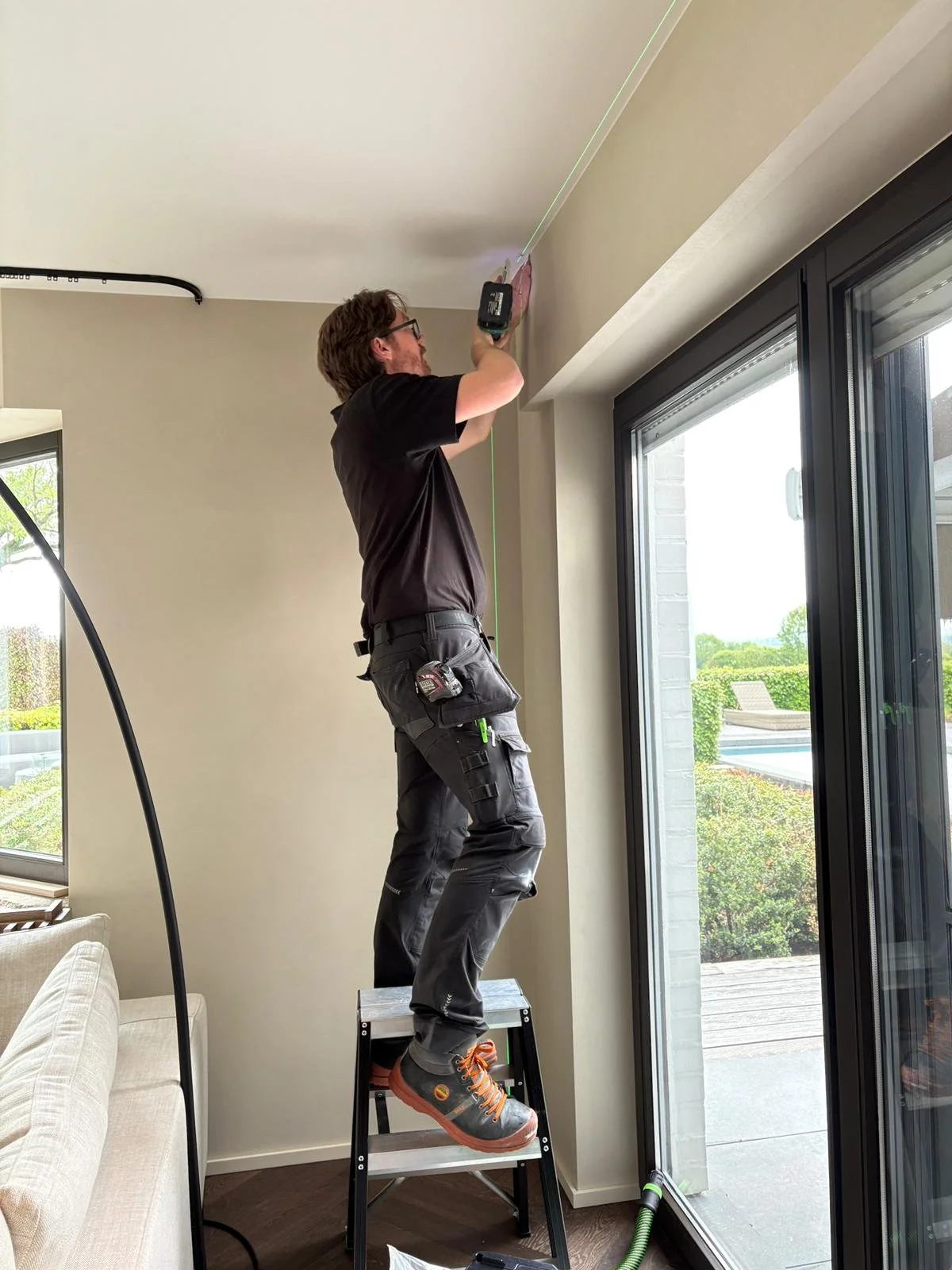 Un homme travaillant à l'intérieur, utilisant une perceuse pour fixer quelque chose au plafond, debout sur un escabeau, près d'une grande fenêtre en verre.
