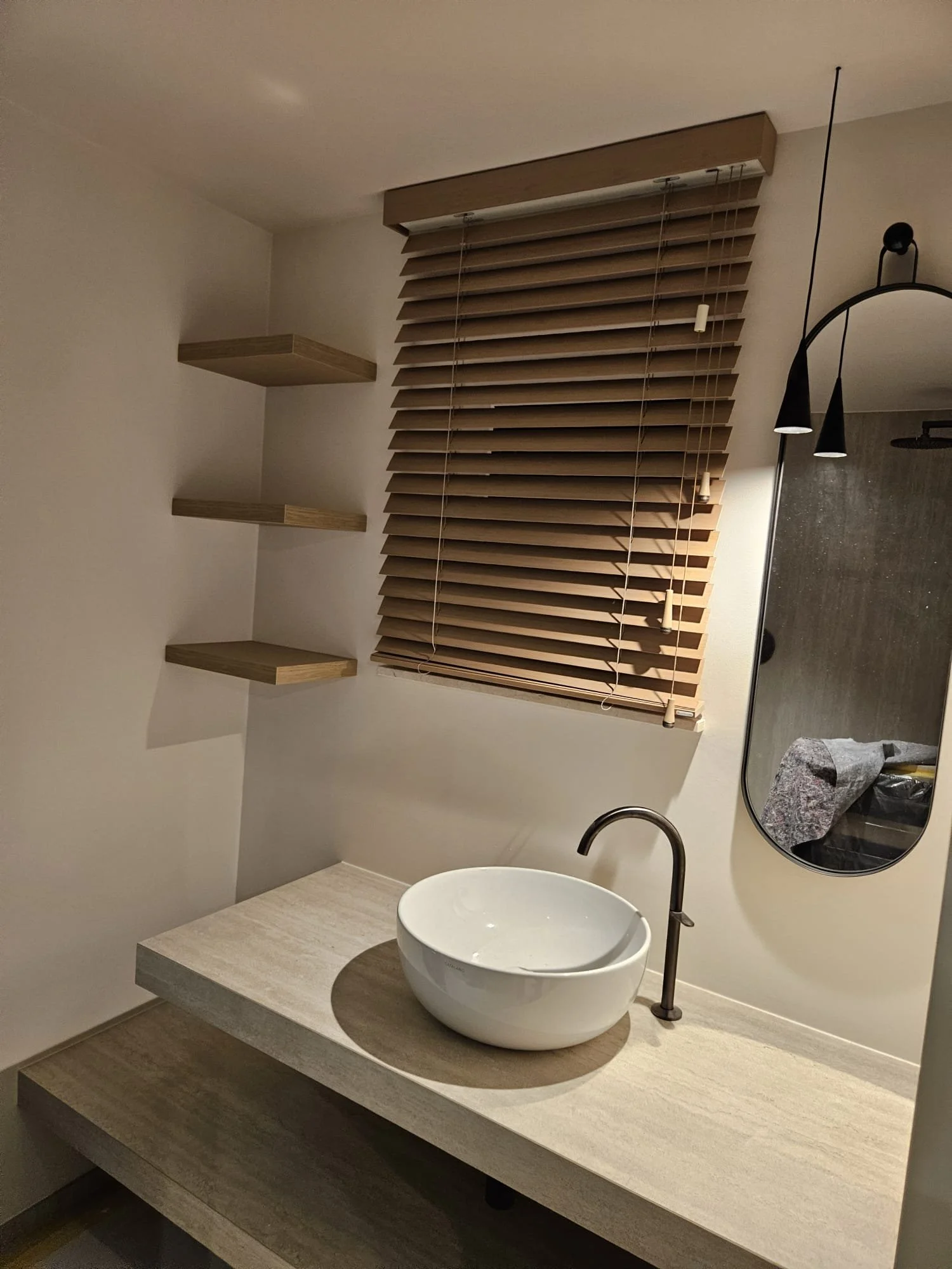Un lavabo rond blanc sur une table en marbre beige avec un robinet noir à côté d'un grand miroir en forme ovale. Sur le mur derrière, trois étagères en bois et une fenêtre avec des stores en bois horizontaux.