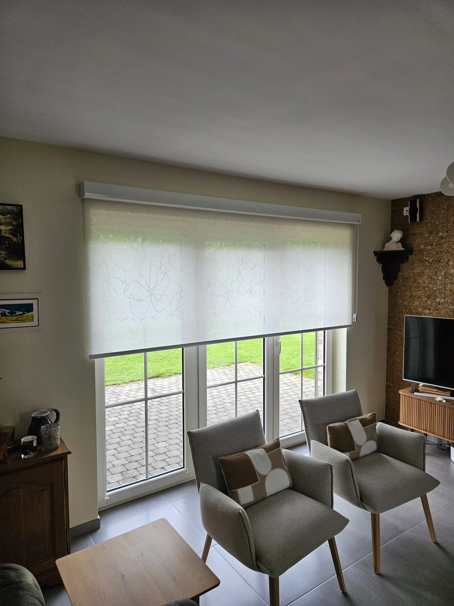 Salon avec vue sur une terrasse extérieure, fenêtres avec rideaux translucides, canapé beige avec coussins, table en bois, télévision, mur décoratif en pierre et une étagère avec une sculpture.