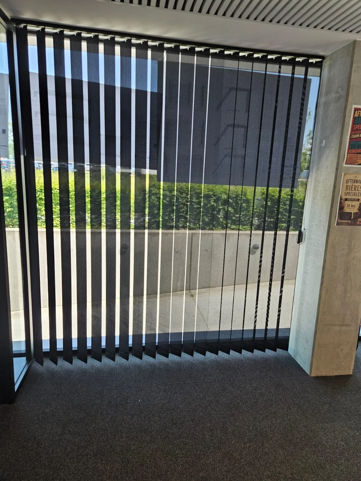 Une vue d'une porte ou fenêtre avec des stores vénitiens en noir, donnant sur un balcon avec une végétation en arrière-plan. À droite, une colonne en béton avec des affiches publicitaires.
