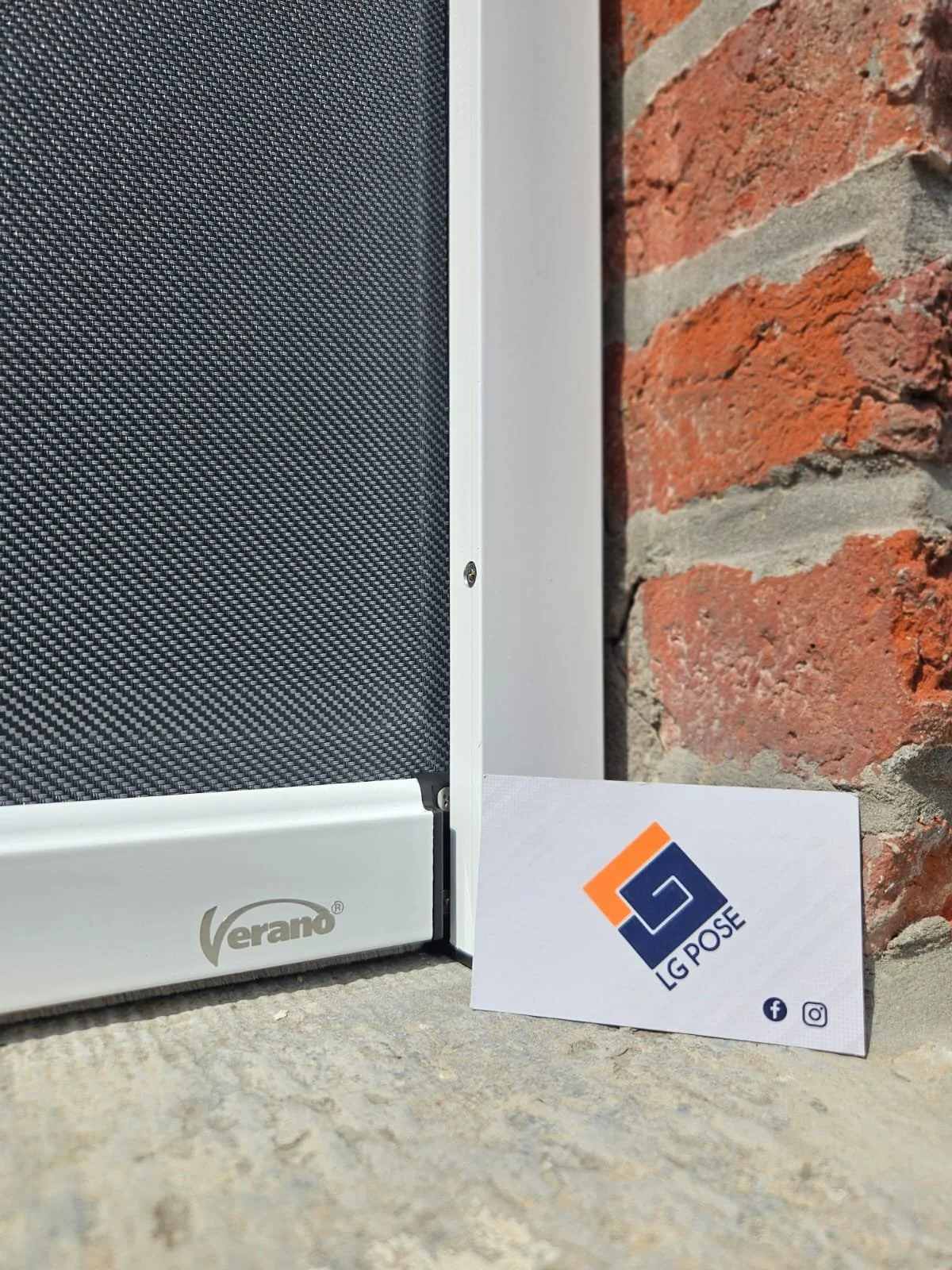 Partie d'une grille ou d'un panneau avec la marque Verano, posé sur un sol en pierre, avec un mur en briques rouges en arrière-plan, et un panneau avec le logo LG POSE à côté.