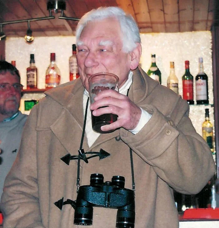Older man in tan coat drinking from a glass at a bar, with binoculars hanging around his neck, and a man in glasses in the background.