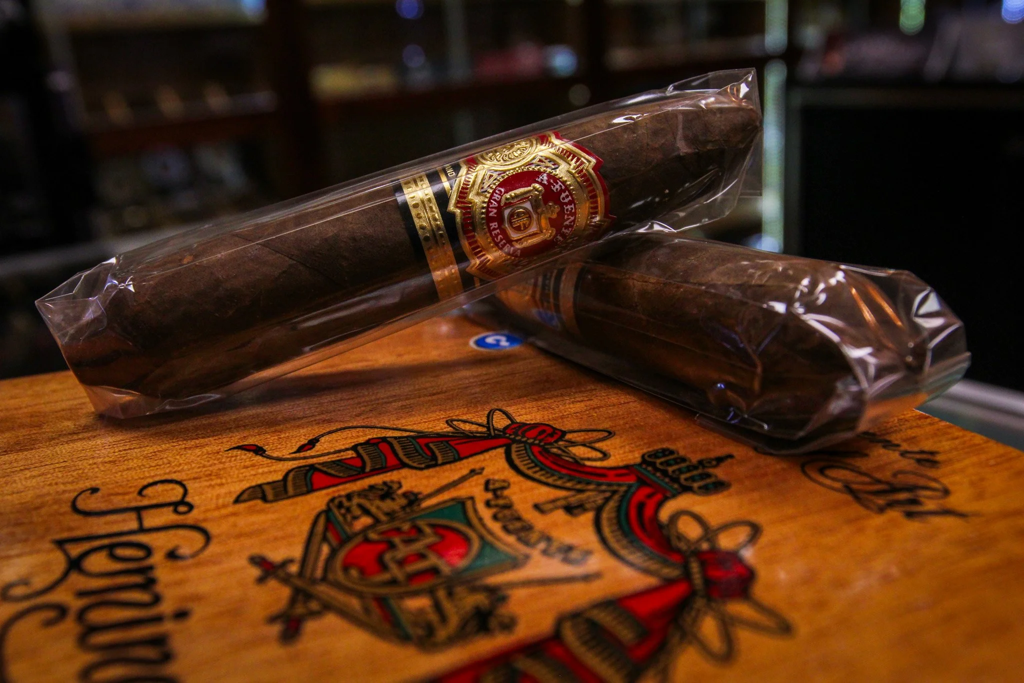 Close-up of a Hemingway cigar inside a clear plastic wrapping on a wooden surface. The cigar has a detailed band with gold, red, black, and white accents. Part of a decorative box or paper with vibrant, colorful graphics is visible below the cigar.