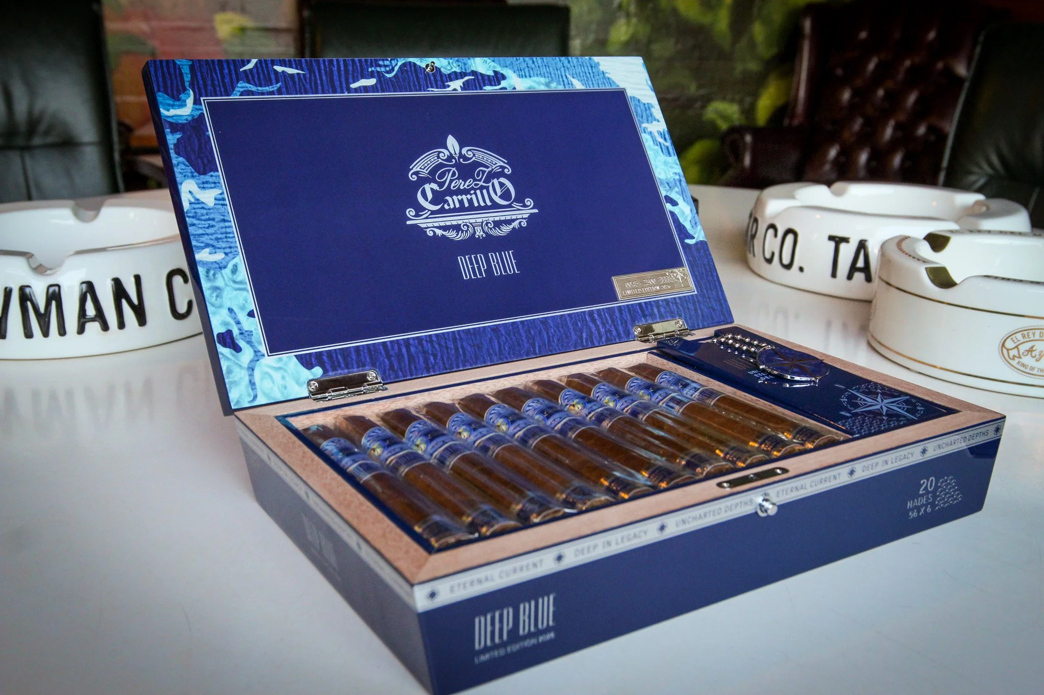 Close-up of a blue and white box of cigars labeled "Deep Blue" by Pere Camillo, with additional cigars and ashtrays nearby on a white surface.