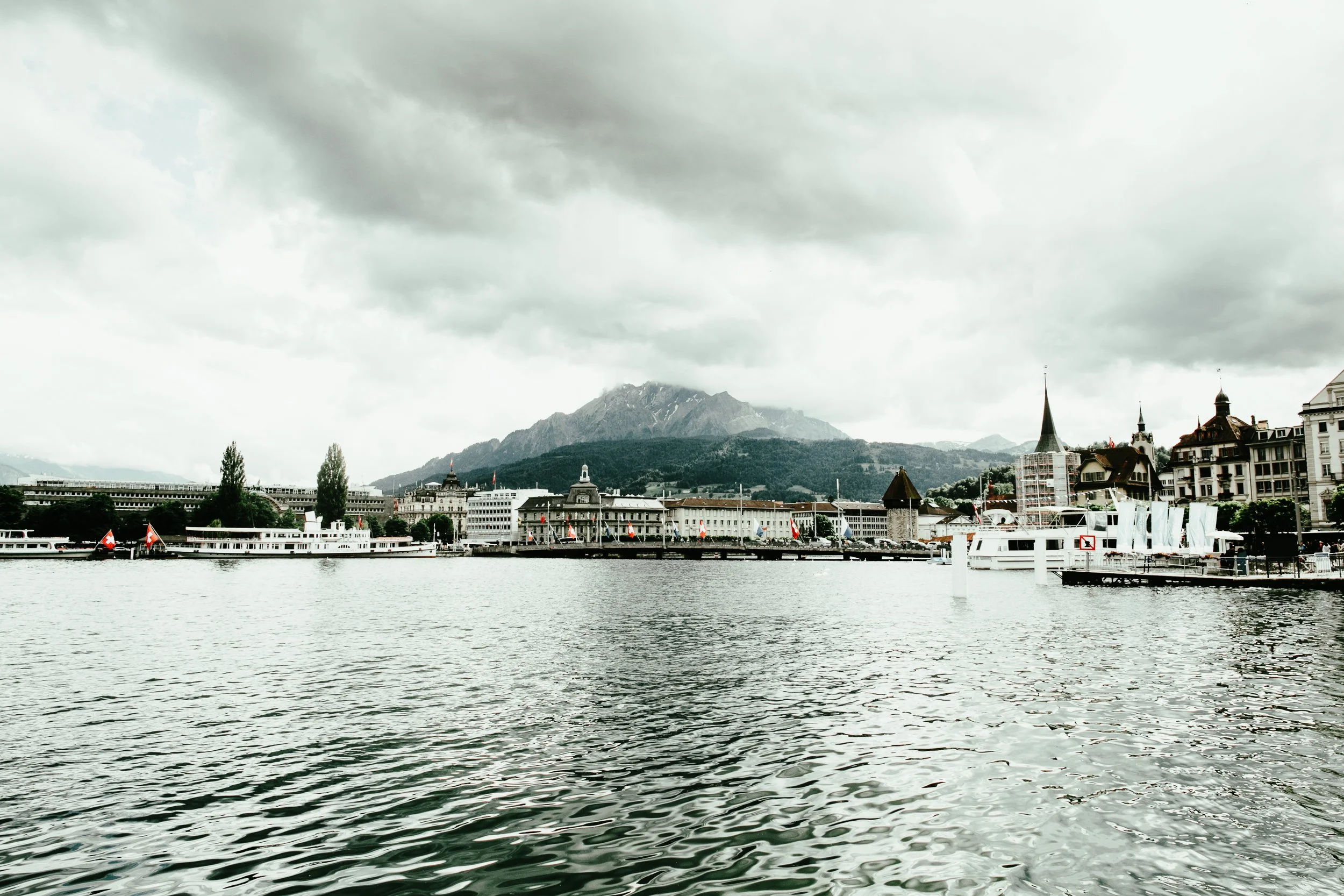 Stadt am Wasser mit Booten, historischen Gebäuden und Bergen im Hintergrund, unter bewölktem Himmel.