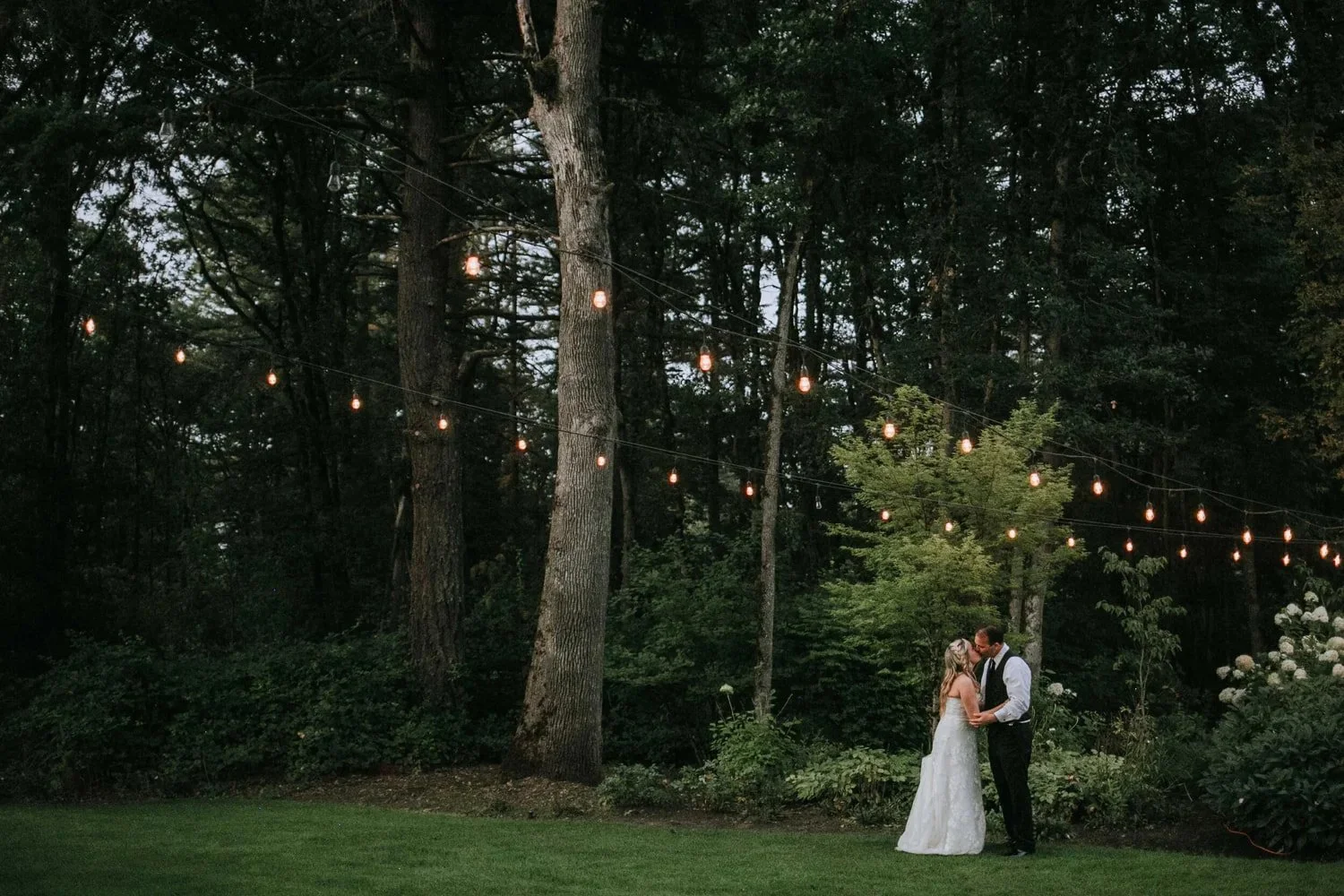 A couple in wedding attire stands together in a lush forest, surrounded by trees and soft natural light.