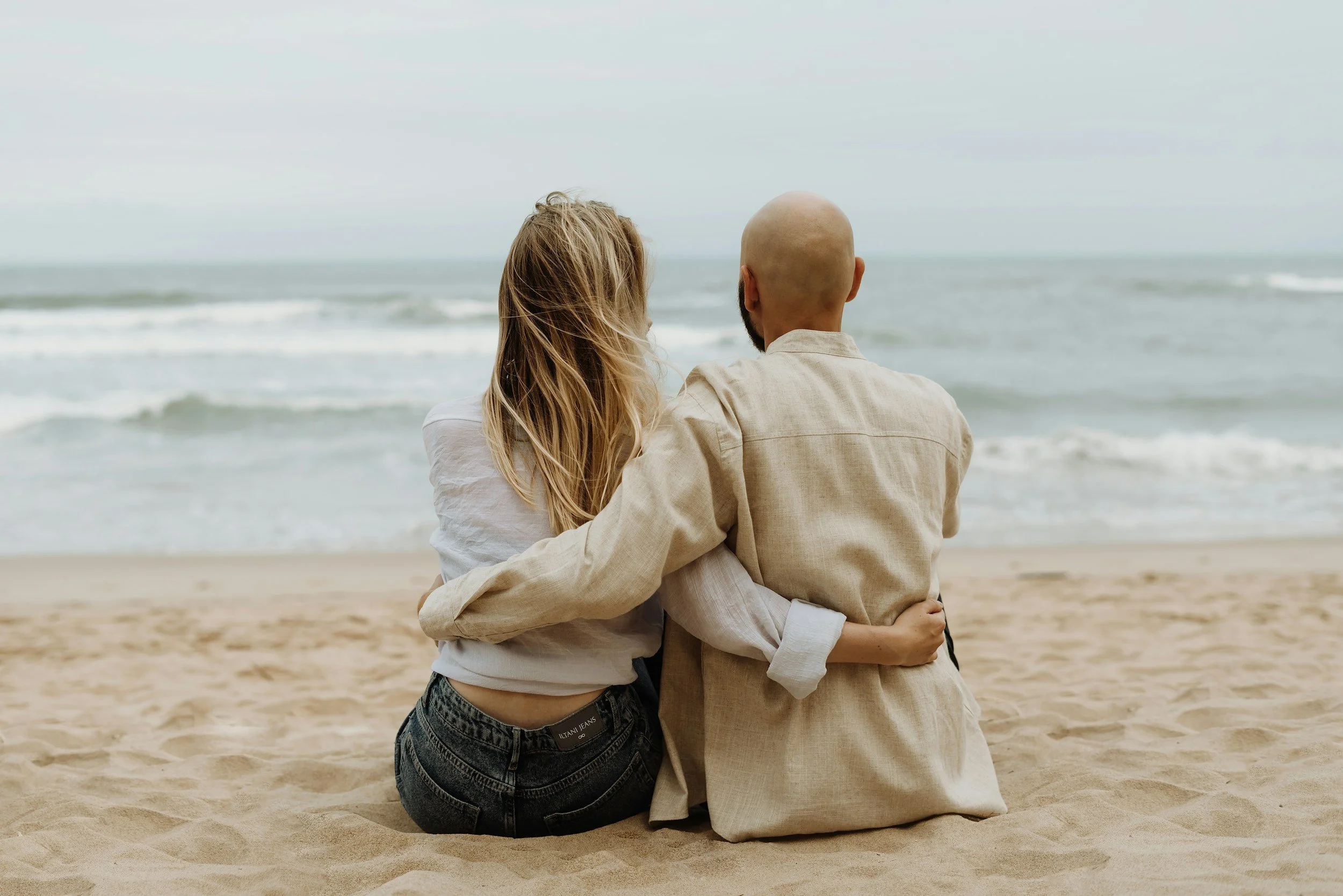 Ein Mann und eine Frau sitzen am Strand, sie umarmen sich und schauen aufs Meer.