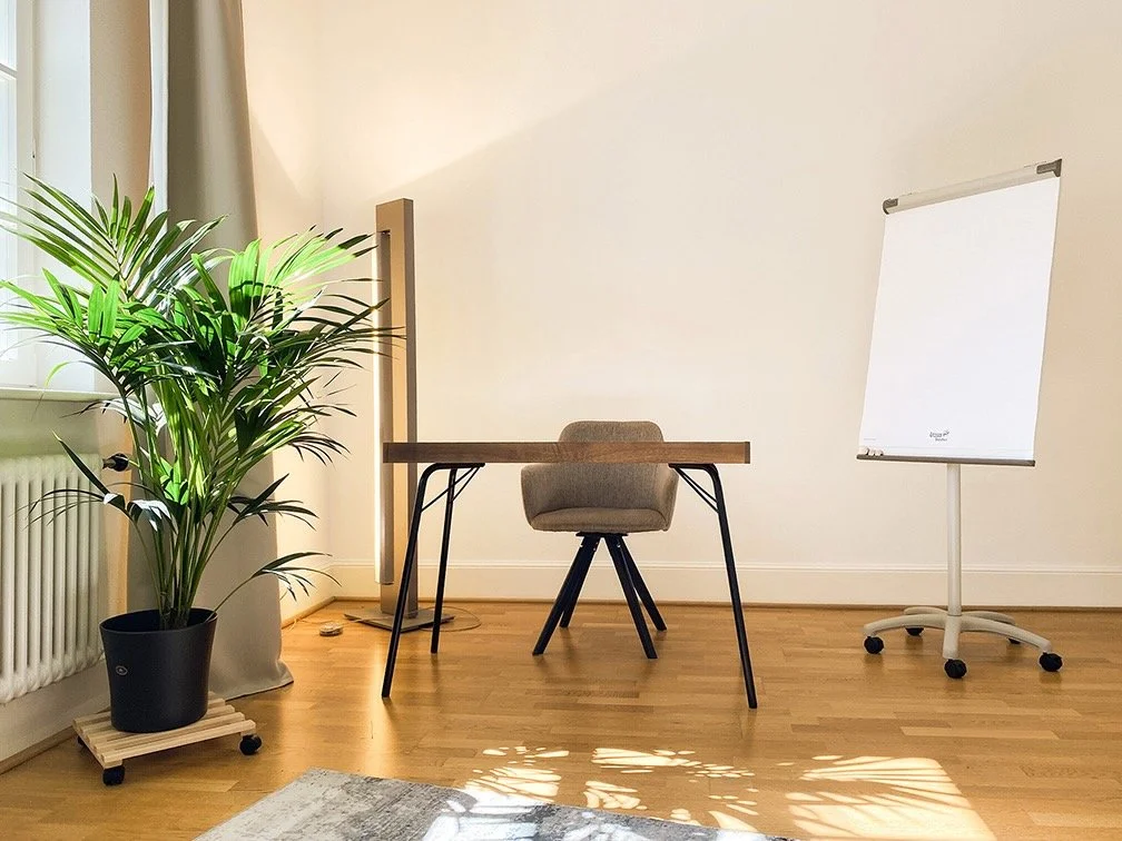 Paartherapieraum in modernem Design mit einem Schreibtisch, einem Stuhl, einem Whiteboard auf einem Ständer, einer Zimmerpflanze und einem Heizkörper in der Ecke.