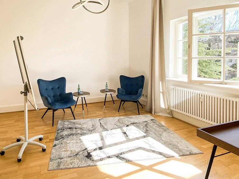 Von Sonnenlicht hell erleuchteter Raum für Paartherapie in Muenchen mit Holzboden, zwei bequemen blauen Sesseln und zwei kleinen Tischen mit Wasserflaschen, eine große Fensteröffnung mit Blick auf Bäume, ein modernes Deckenlicht.