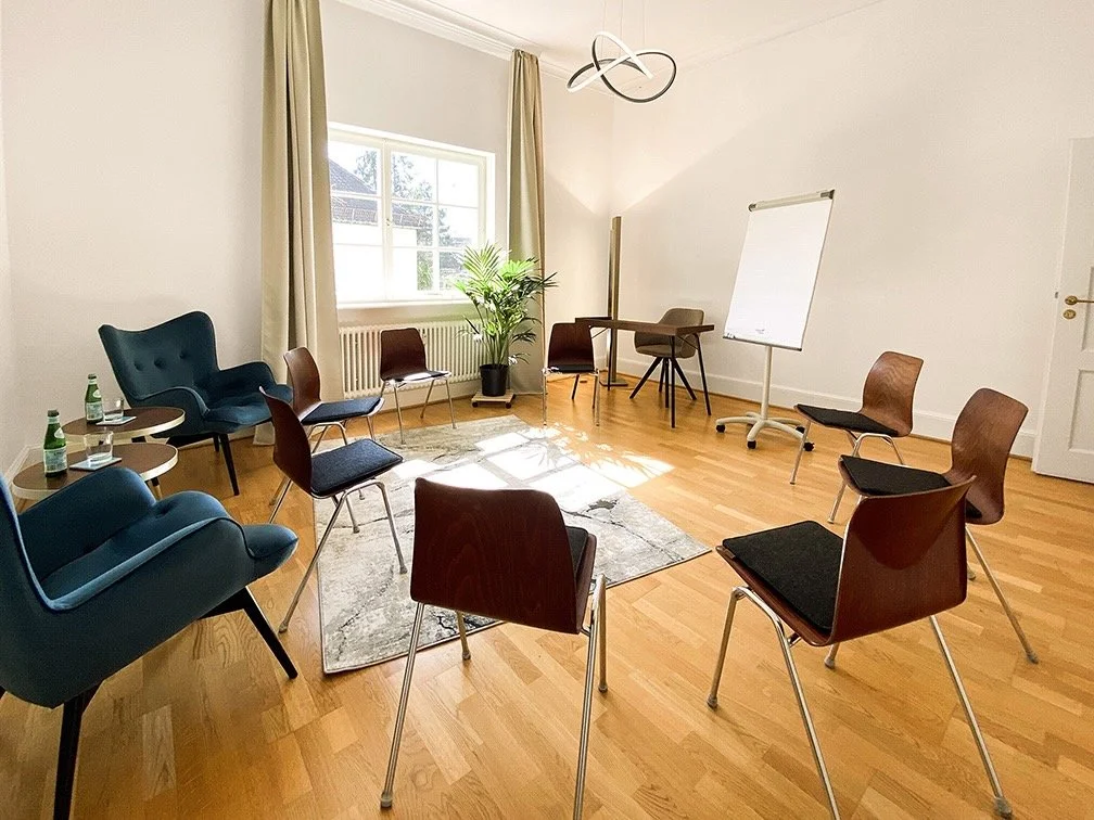 Therapieraum von Dr. Ildi Sarosi mit Stuhlkreis, Pflanzen, Whiteboard, Tisch, und Sonnenlicht durch das Fenster.