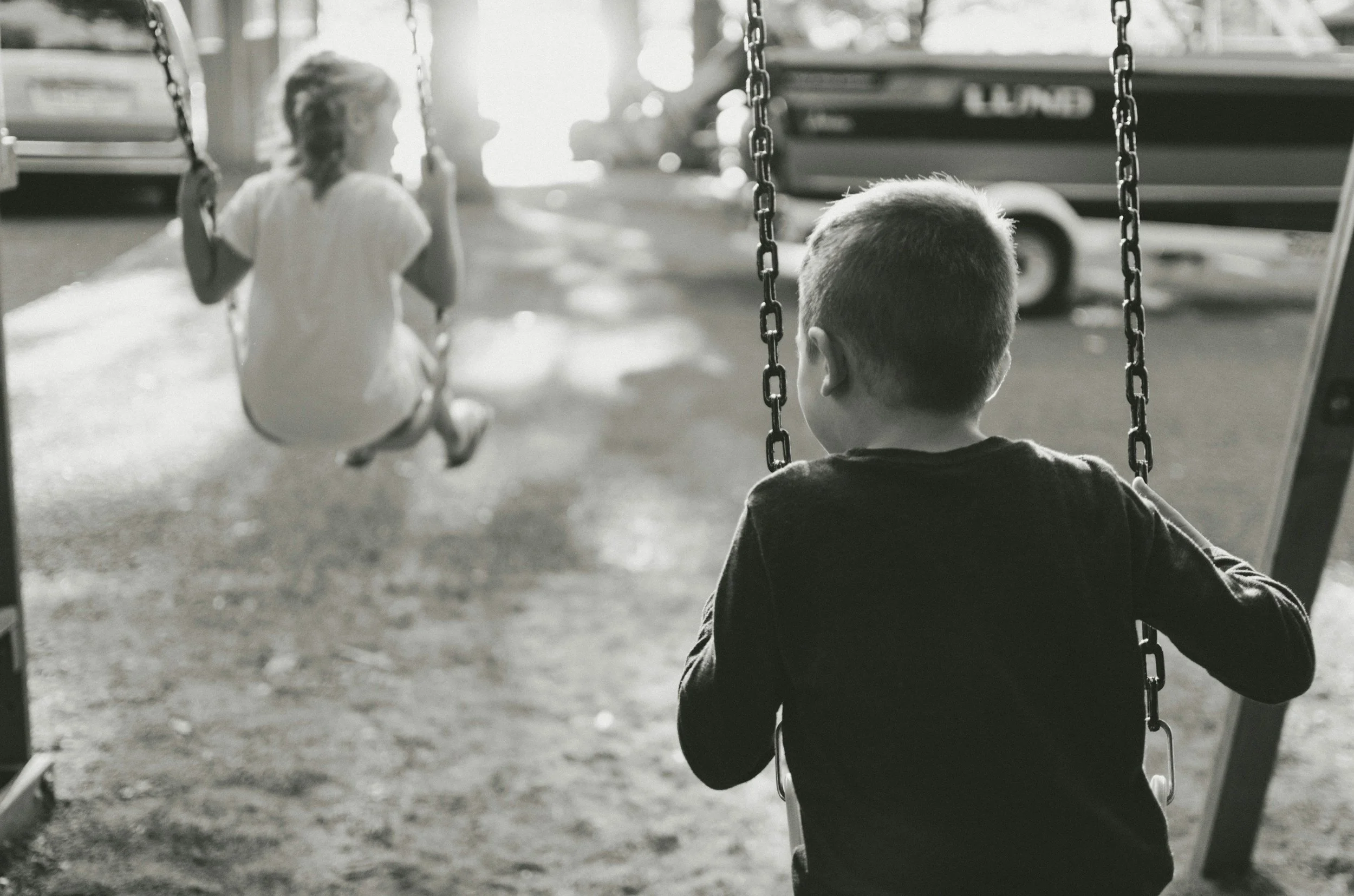 Un garçon assis sur une balançoire regarde une fille qui joue, dans un parc, en noir et blanc.