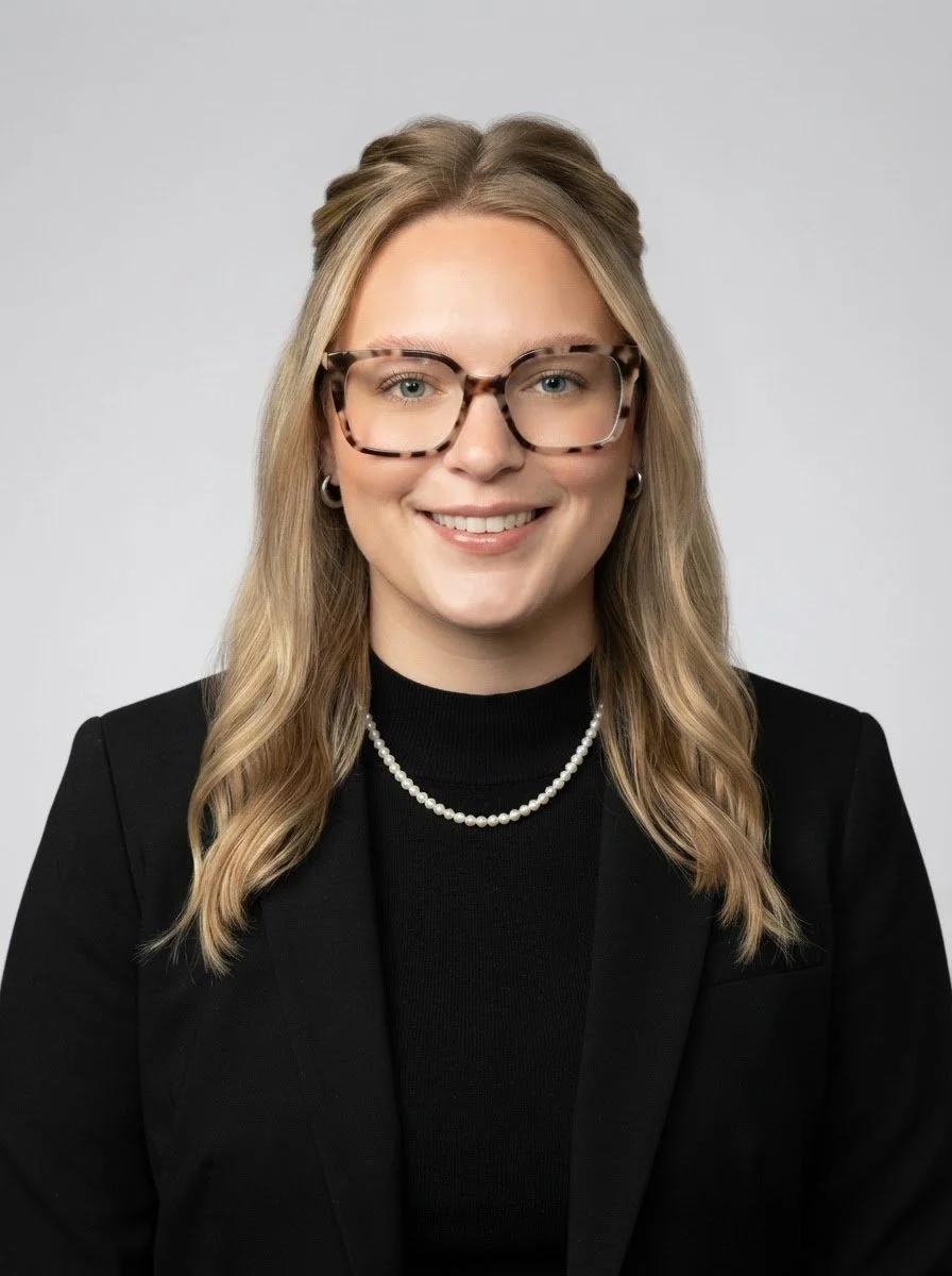 Portrait d'une femme souriante portant des lunettes à motif tacheté, une robe noire, un collier de perles et des boucles d'oreilles en argent, contre un fond gris clair.