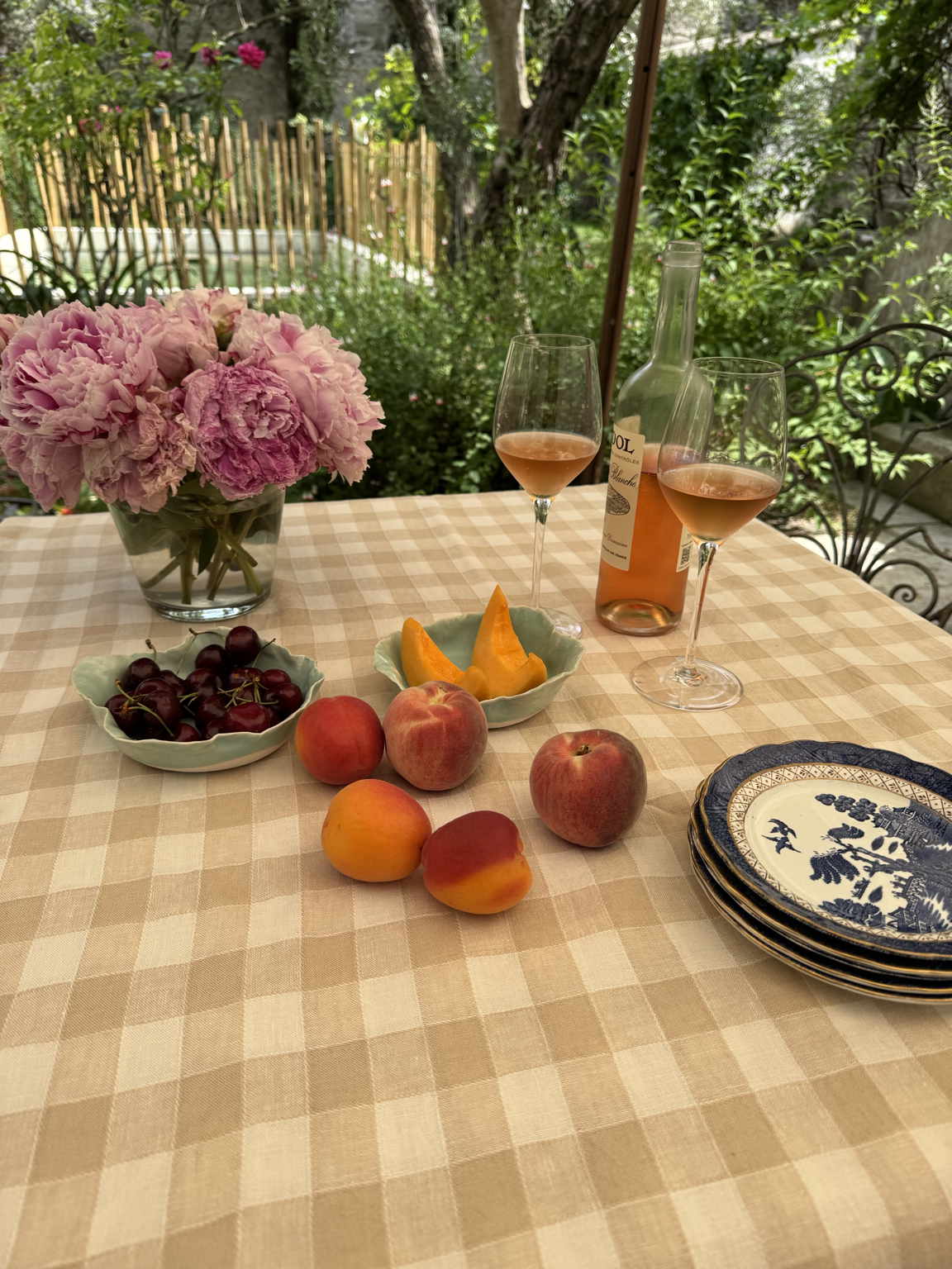 A table set outside with pink peony flowers in a vase, a bowl of cherries, peaches, and apricots, two glasses of rosé wine, a wine bottle, and a stack of blue and white plates. The background features trees, plants, and sunlight.