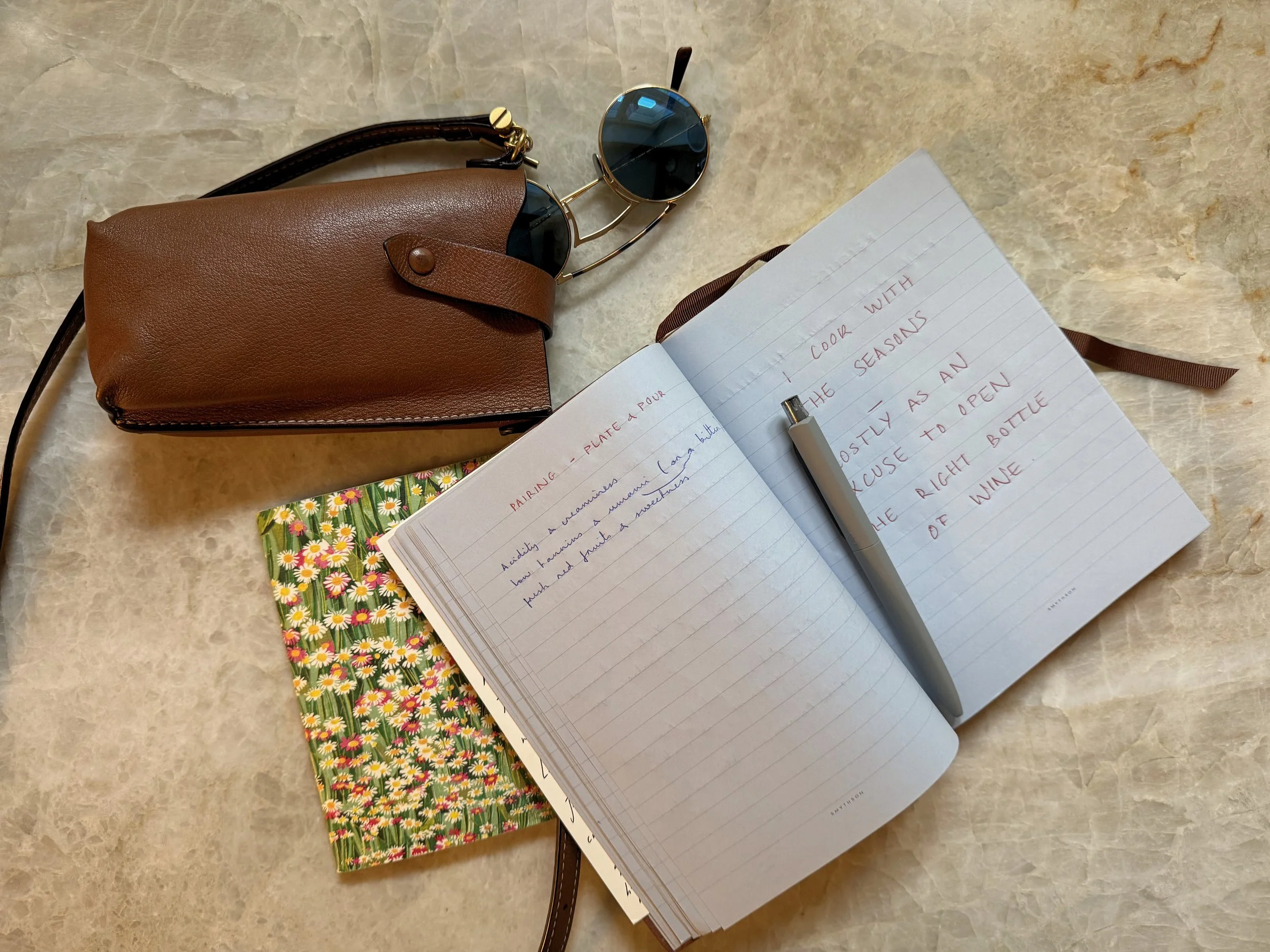 A collection of personal items on a marble surface, including a brown leather bag, black sunglasses with round lenses, a lined notebook with handwritten notes, a pen, a floral-patterned envelope, and a piece of paper with handwritten text.