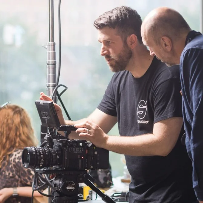 Two men working with professional filming equipment inside a room with large windows, one man with a beard and dark hair wearing a black shirt, the other bald and wearing a dark jacket, both focused on the camera setup.