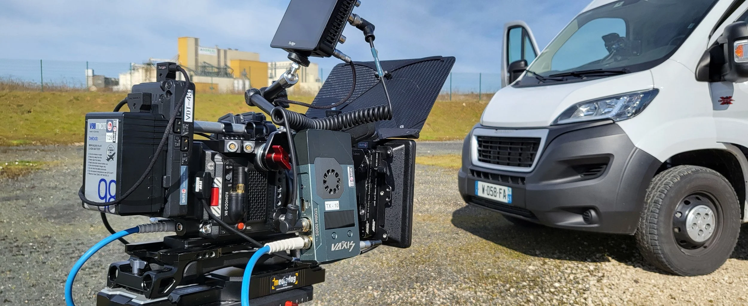 Close-up of a professional video camera mounted on a rig facing a white van parked outdoors, with factory buildings in the background.