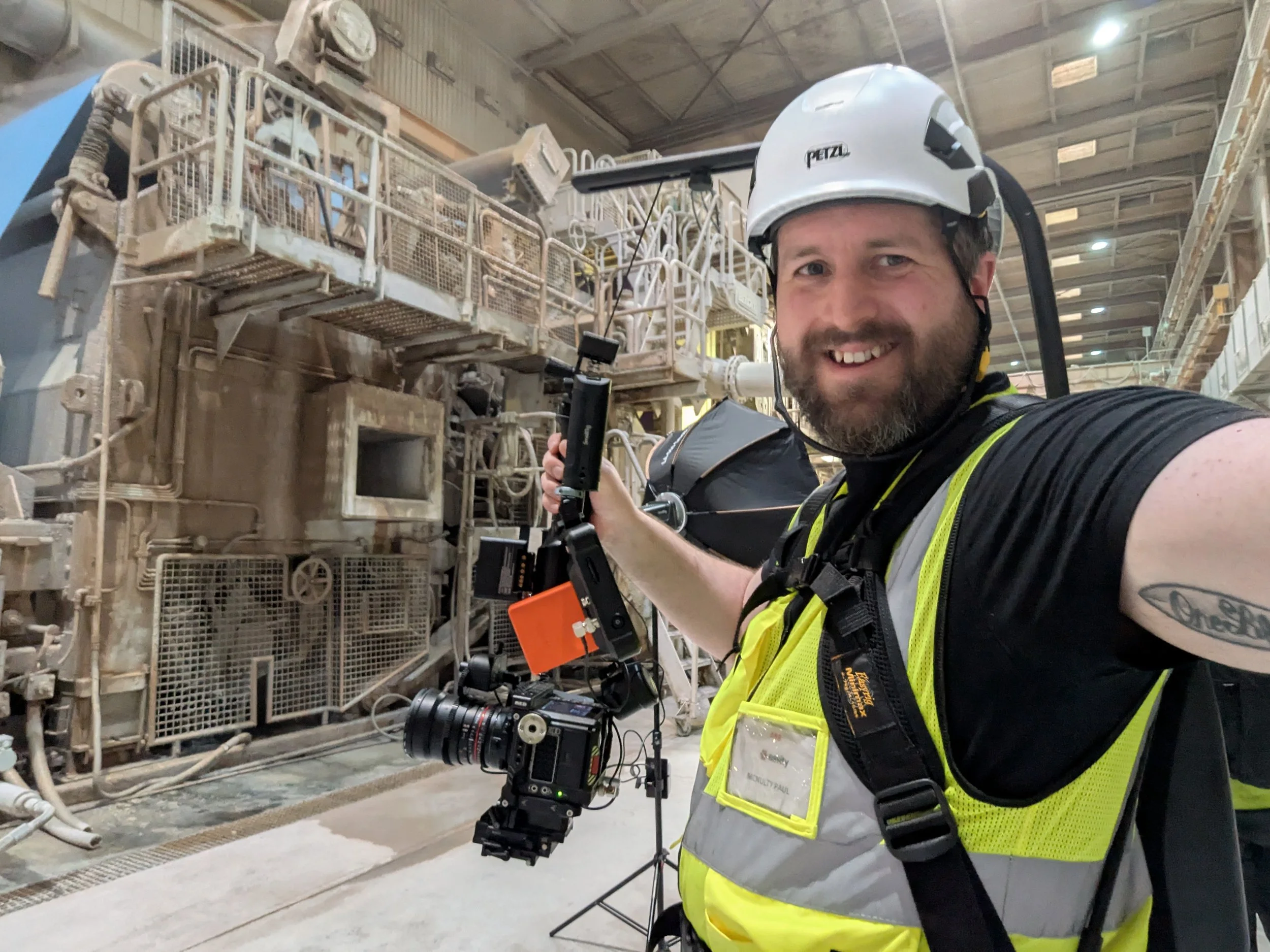 A man with a beard, wearing a safety helmet and vest, is taking a selfie in an industrial setting with complex machinery and equipment in the background, holding a camera on a stabilizer.