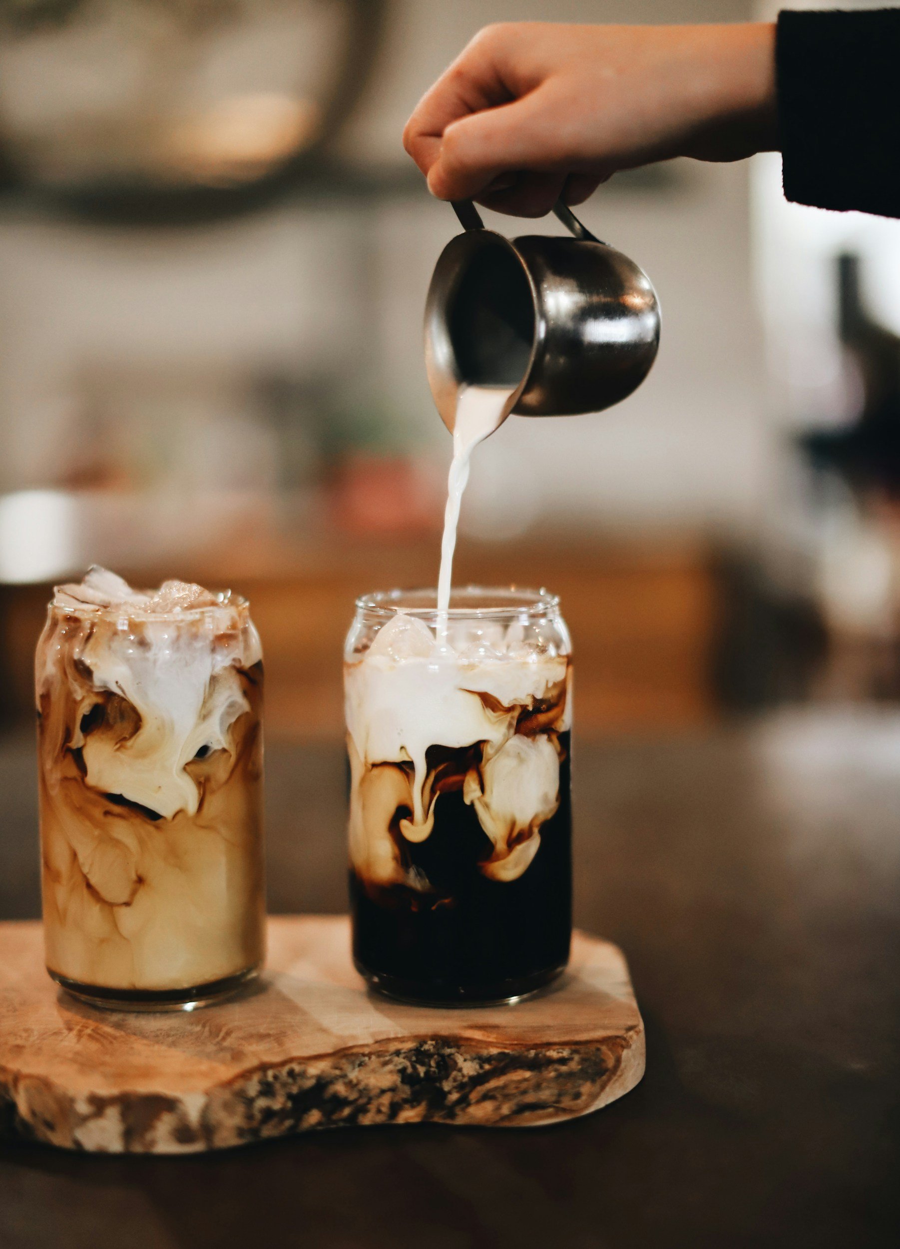 Twee glazen met iced coffee met melk en chocolade, op een houten plank, de ene met een zwarte vloeistof, de andere met melk, een lepel met melk wordt in de zwarte iced coffee geschonken.