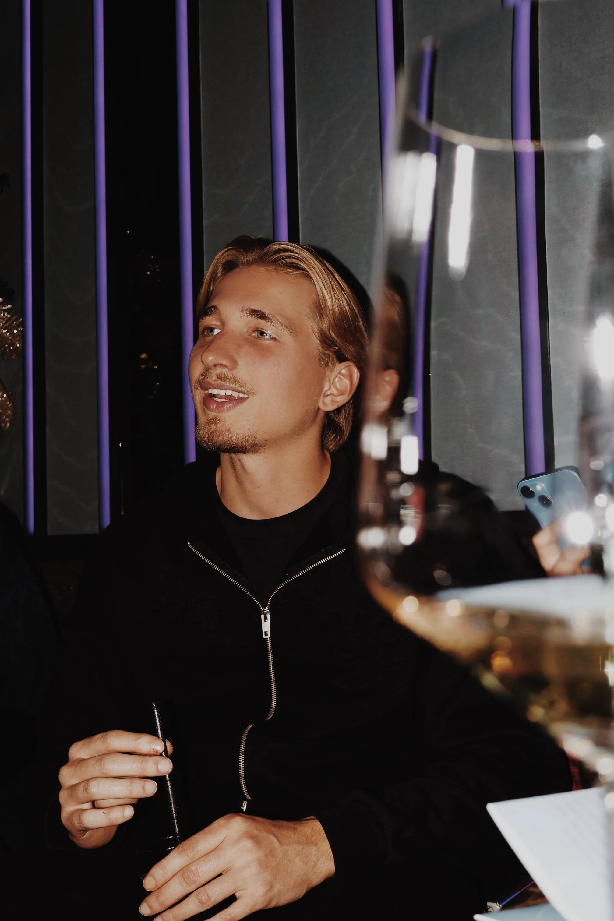 Een jonge man met blond haar en een baard die aan een feest of diner deelneemt, zittend aan een tafel met een glas wijn en een sigaret, omringd door andere gasten.