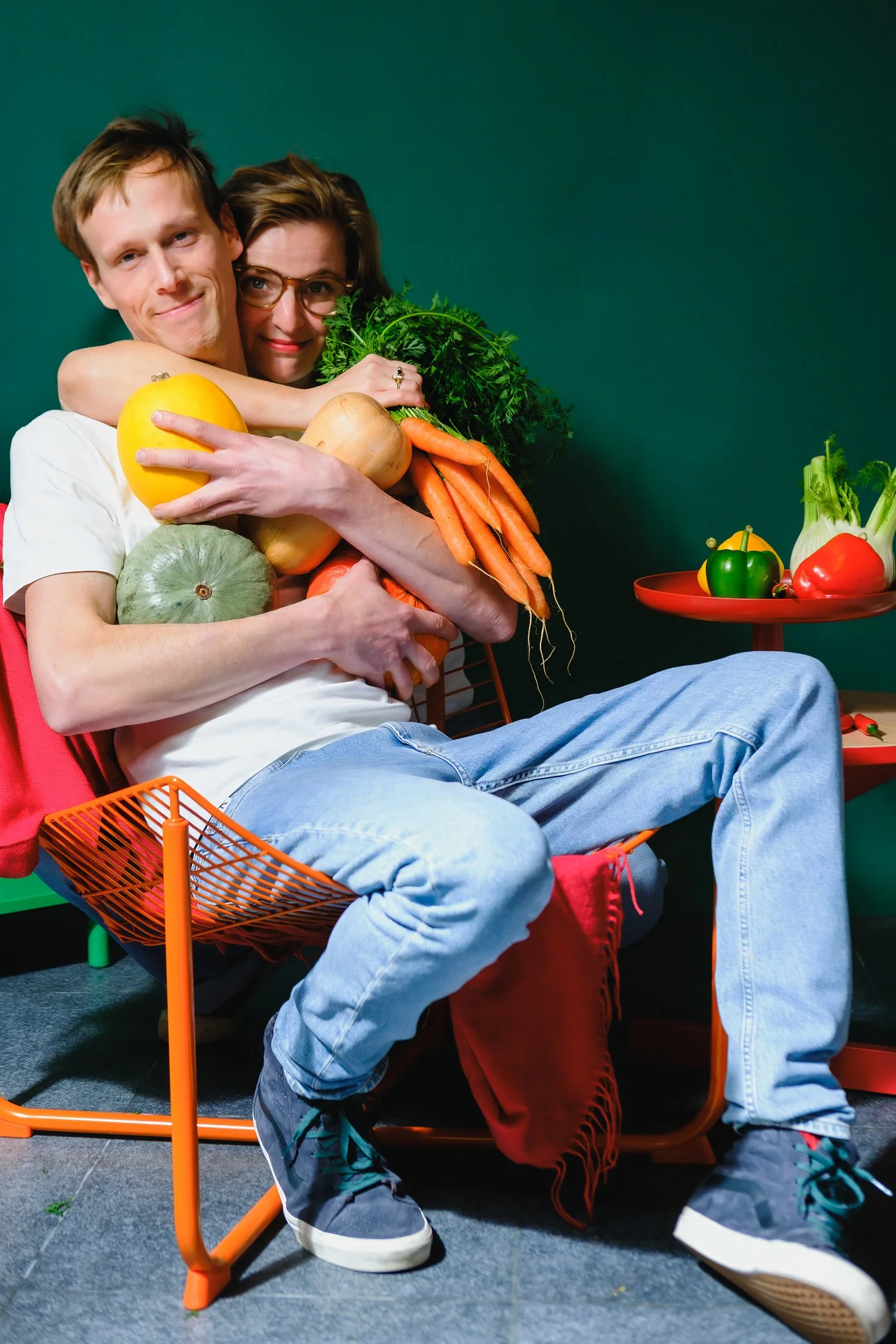 Een man en vrouw omhelzen elkaar terwijl ze een grote hoeveelheid groenten vasthouden, zoals wortels, courgette, paprika, pompoen en peterselie, met een groene achtergrond.