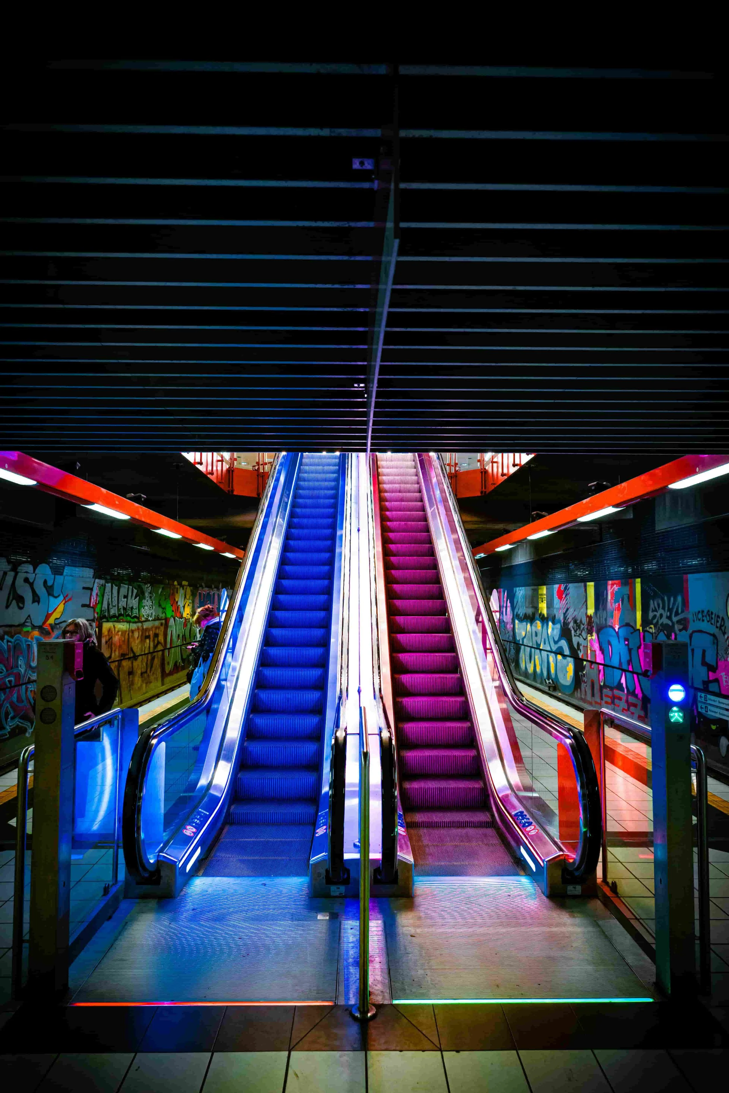 Due scale mobili di colori blu e rosa in un centro commerciale sotto un soffitto nero con luci a LED colorate, pareti decorate con graffiti.