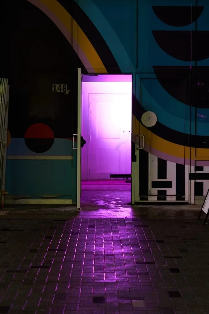 Porta aperta con luce rosa intensa oltre, riflesso rosa sul pavimento di mattonelle.