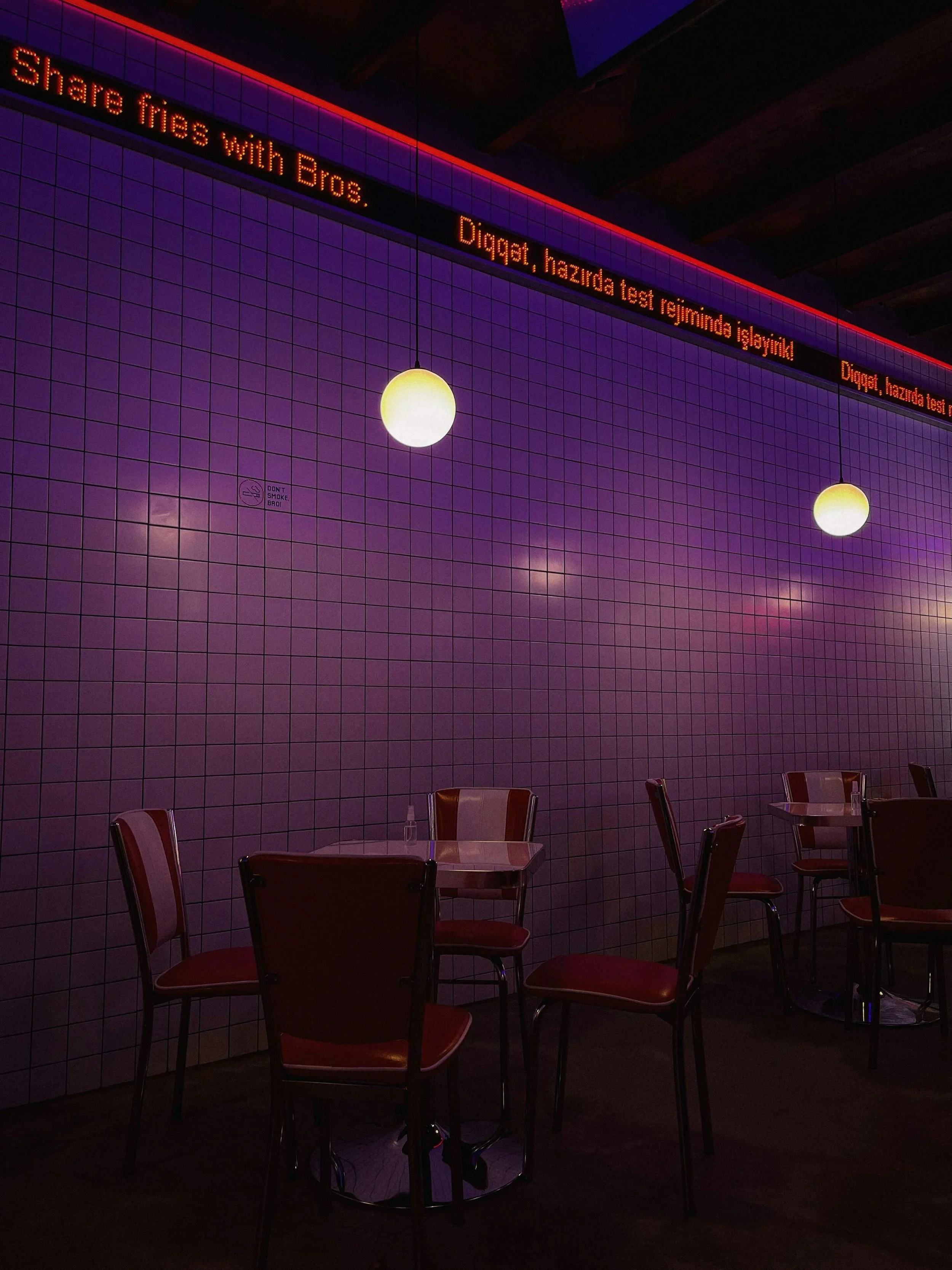 Interno di un bar o ristorante con pareti rivestite di piastrelle rosa e sedie rosse attorno a tavoli rotondi, illuminato da luci a sfera e una striscia di luci LED sul soffitto. Un cartello luminoso mostra testo in lingua turca.