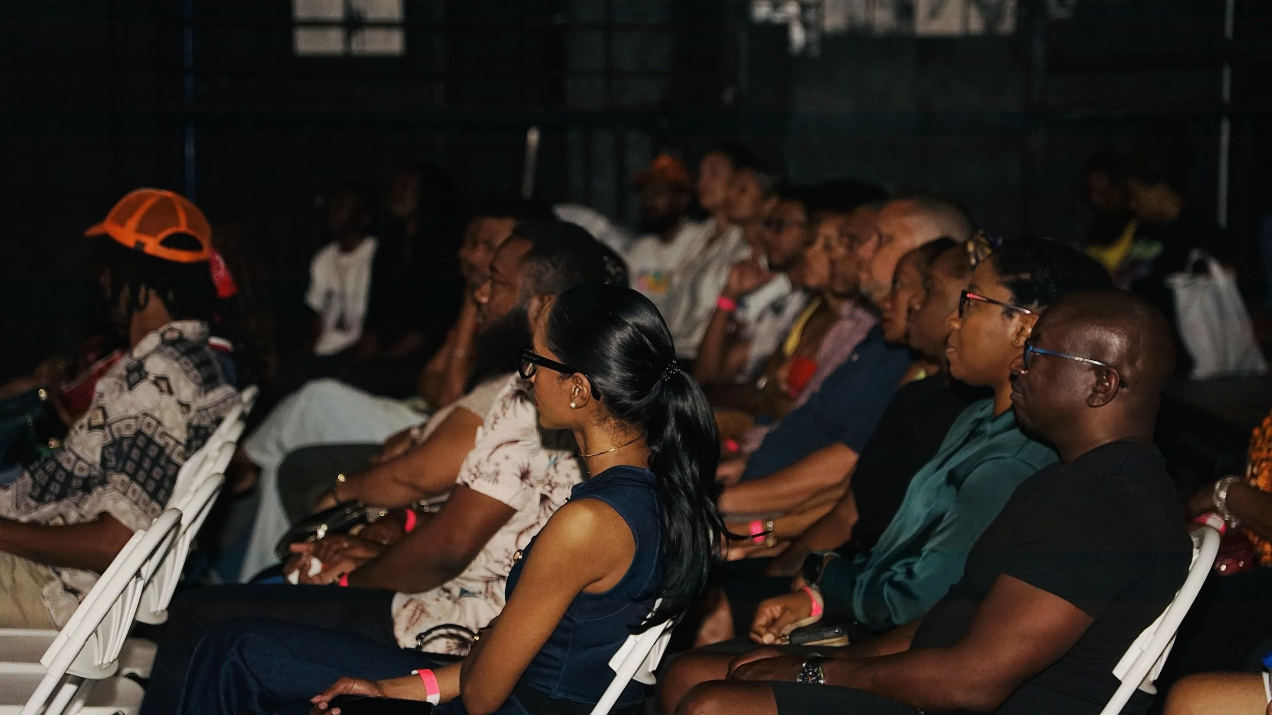Audience seated in a darkened auditorium, listening to a presentation or event, with a woman wearing glasses in the foreground and some audience members wearing wristbands.
