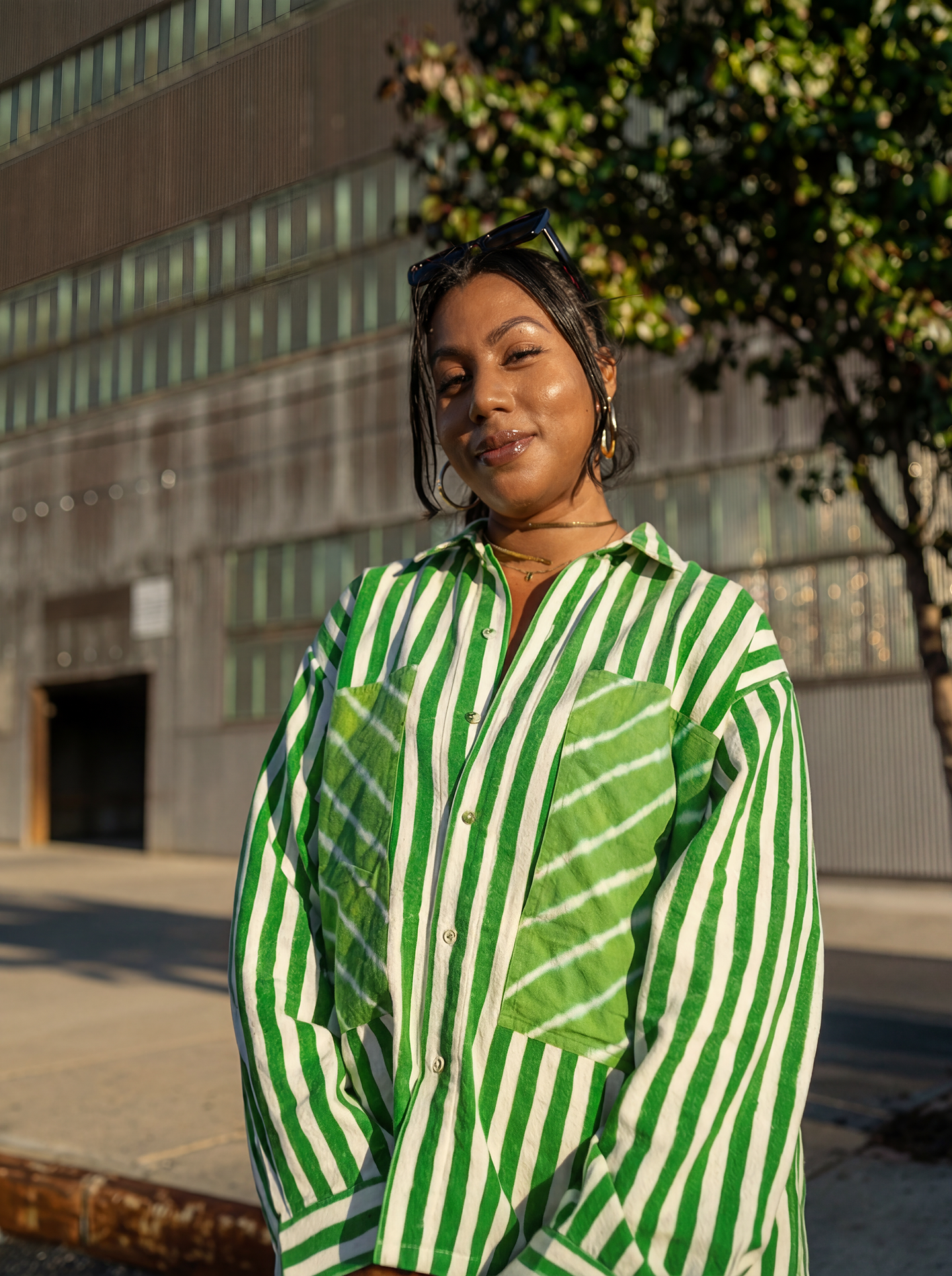 Young woman with dark hair and gold jewelry wearing a green and white striped shirt, standing outdoors near a modern building and trees.