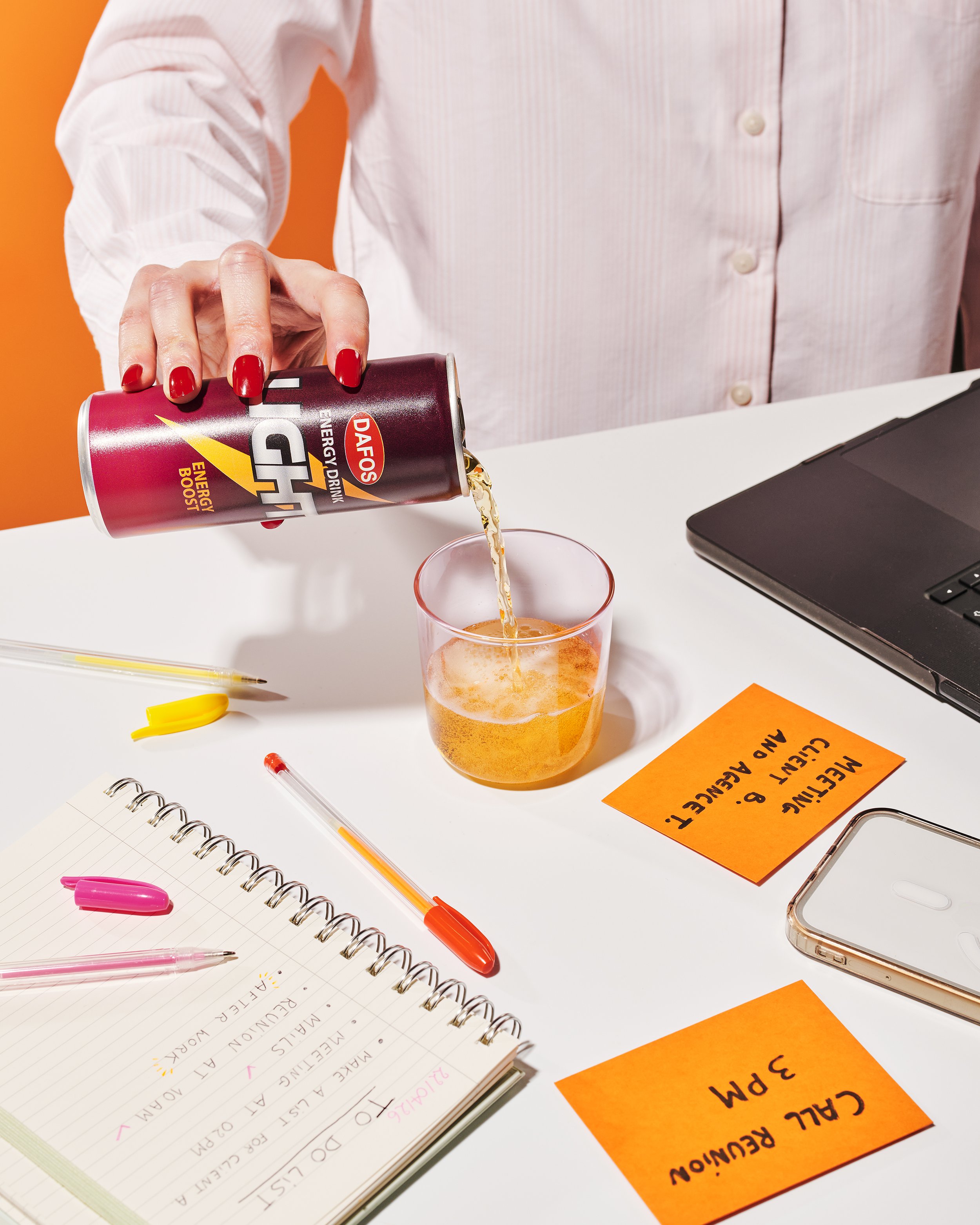 Une personne verse une boisson énergisante sur un verre. Sur la table, il y a un carnet avec des notes, des stylos colorés, des post-it oranges avec d'autres notes, un ordinateur portable et un téléphone portable.