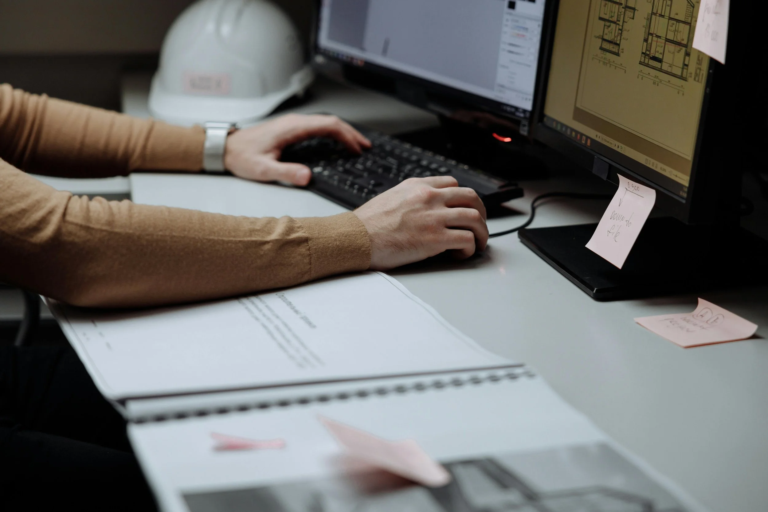 Une personne travaillant sur un ordinateur avec deux écrans, portant un bracelet métallique. Une casquette blanche est posée à côté de l'écran, et plusieurs papiers avec notes écrites sont visibles sur le bureau.