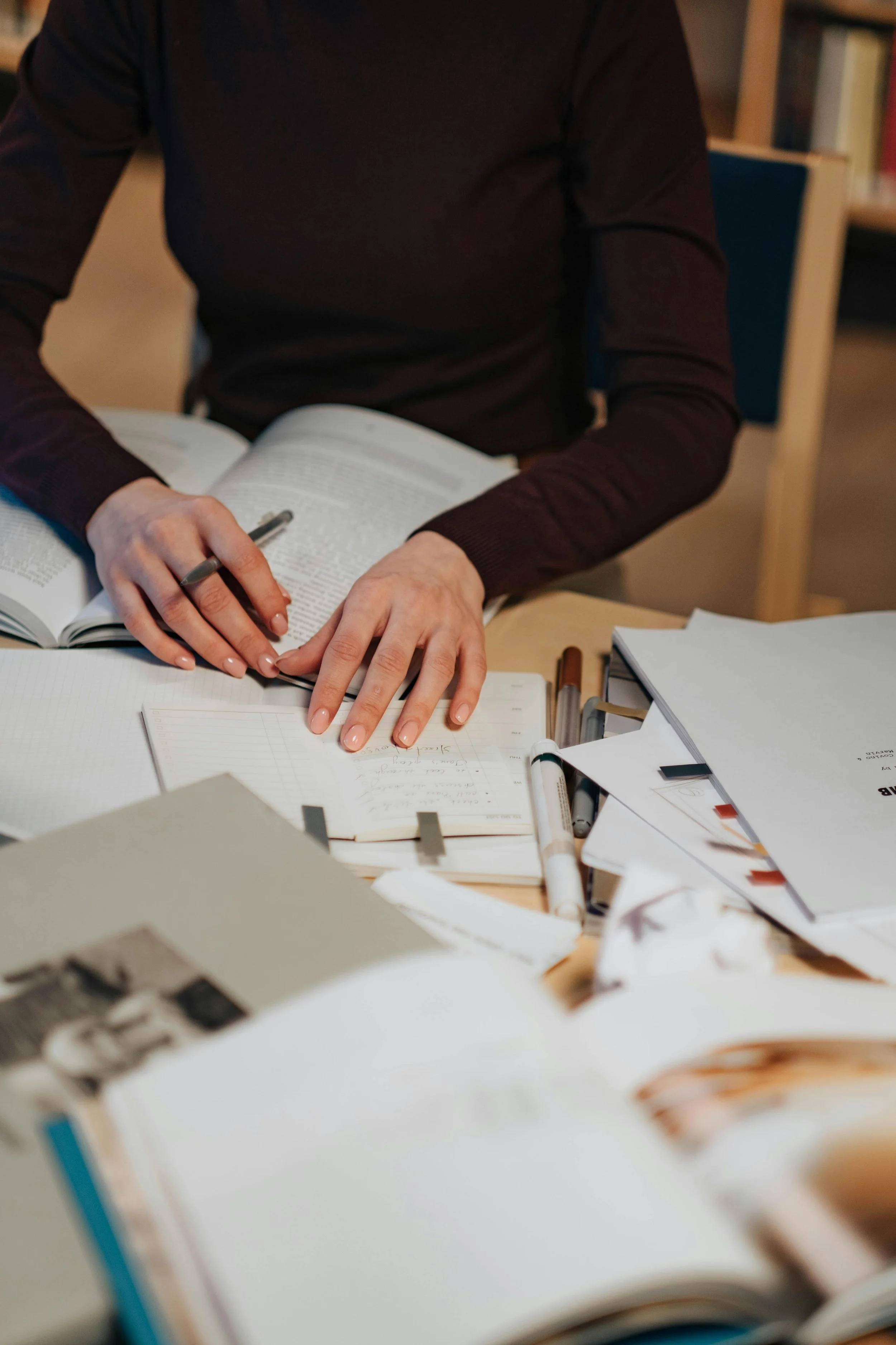 Une personne en sweater marron écrit dans un carnet ouvert sur une table encombrée de papiers, stylos et marqueurs, dans un environnement de bibliothèque ou de bureau.