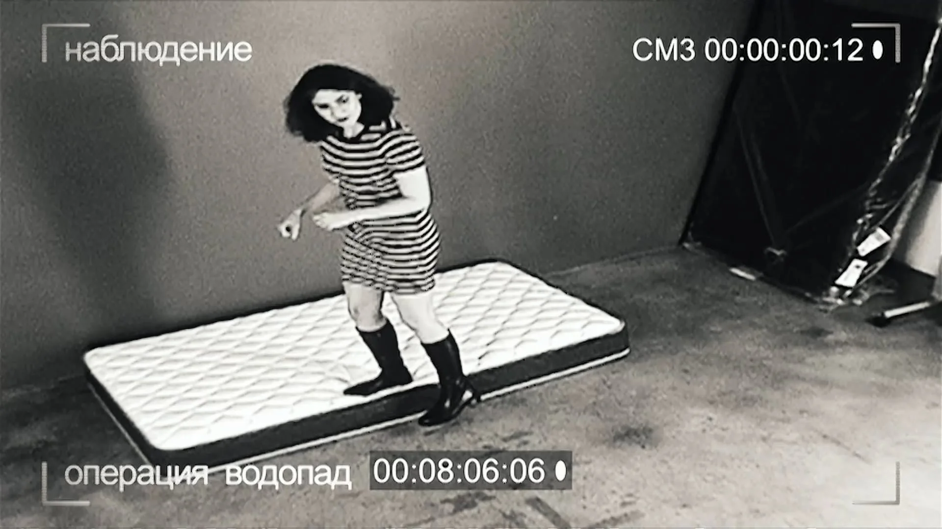 A woman in a striped dress and knee-high boots is standing on a mattress in a room with a dark wall, while looking down and smiling. There is a stacked drawing board or folding table on the right side of the image.