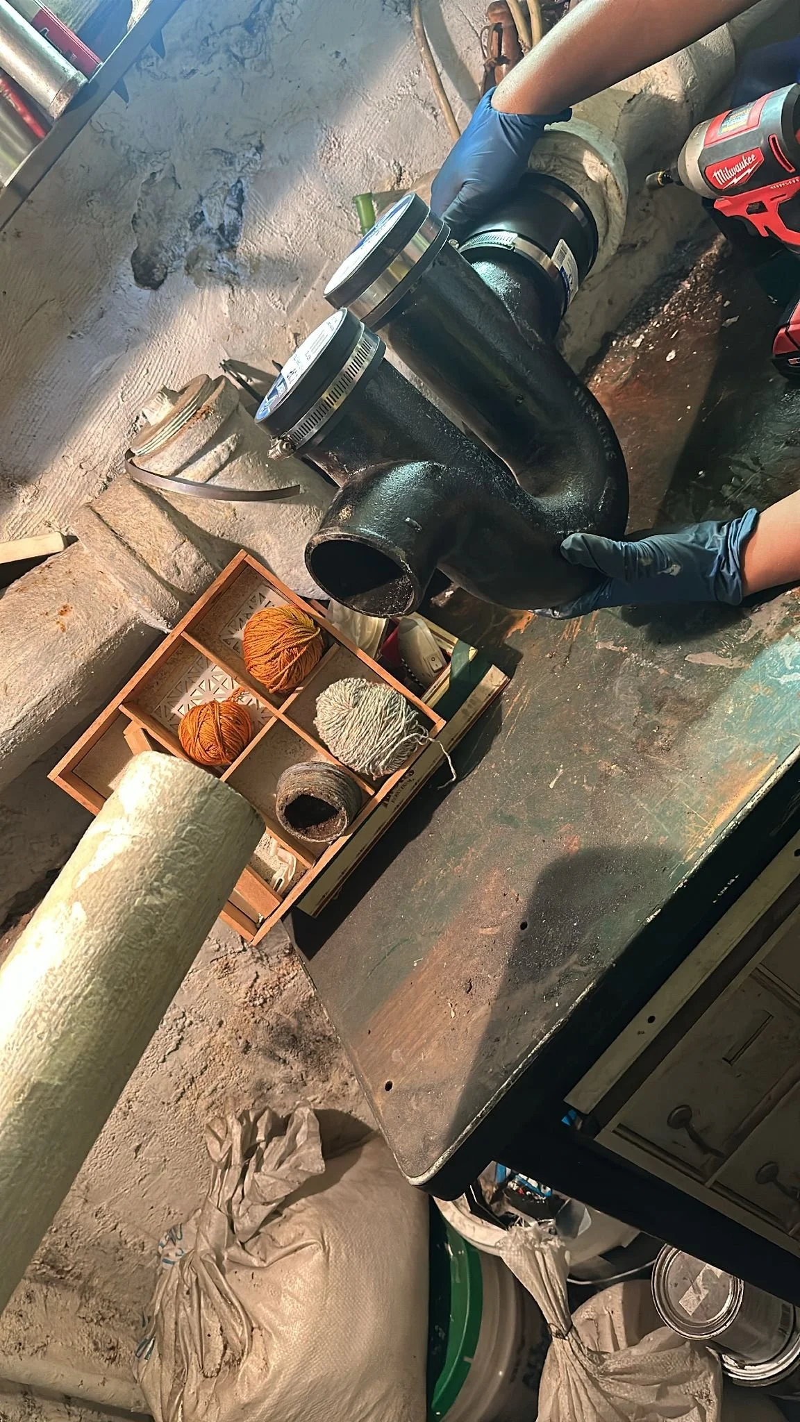 A person in a blue glove installing a black pipe fitting onto a concrete pipe using a power drill. The work surface is cluttered with yarn, tape, cans, and other tools and supplies.