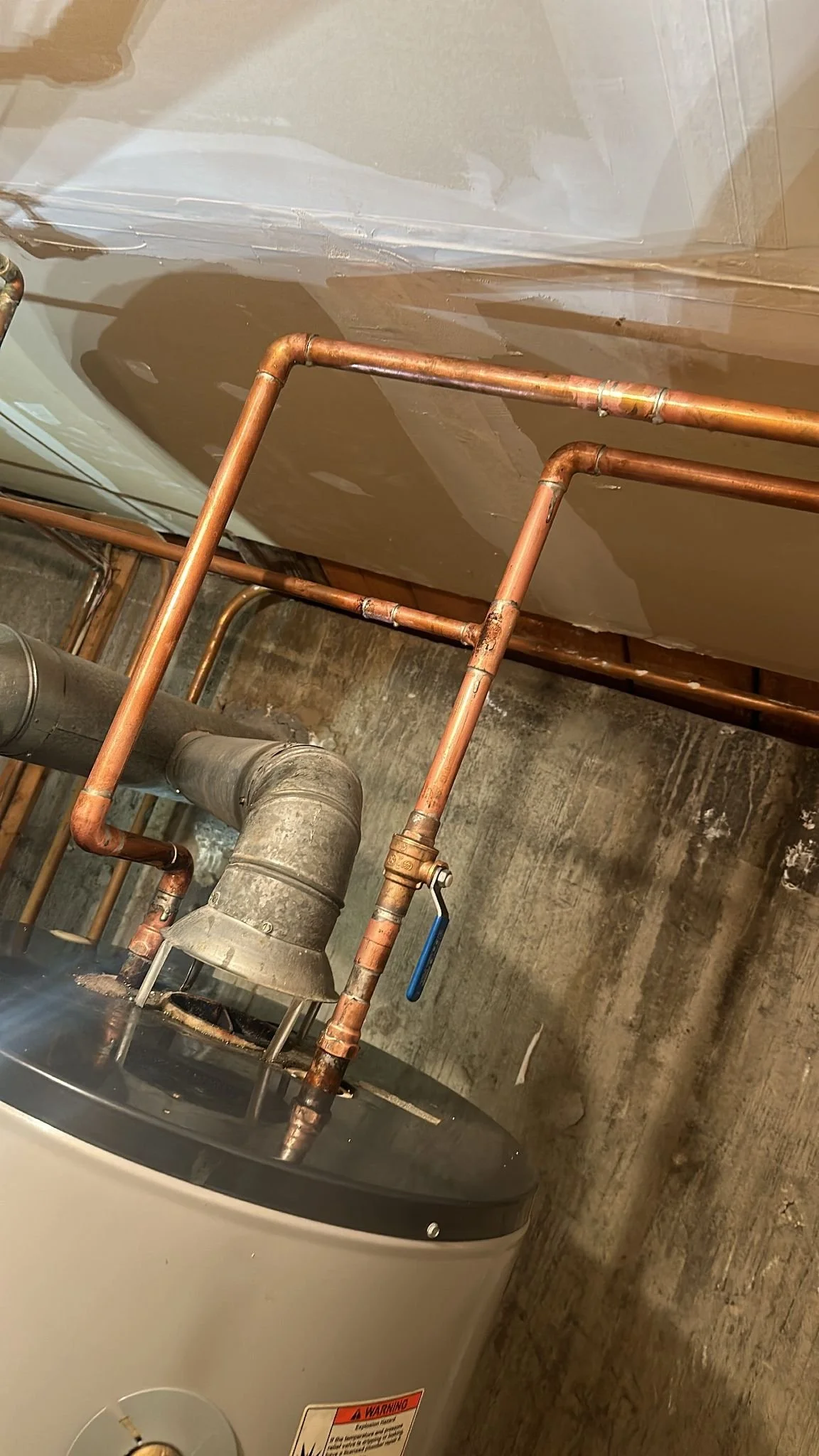 A washing machine connected to orange copper water pipes in a utility room with a rough concrete floor and a plastic sheet taped along the wall.
