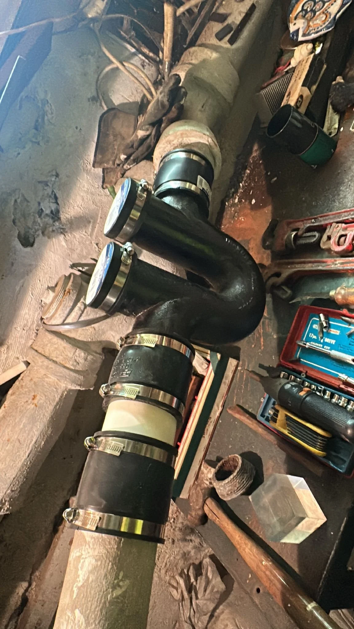 Close-up of a large black pipe with multiple metal clamps attached, mounted horizontally and connected to a beige cylindrical component. The workspace shows tools, a red toolbox, and other mechanical parts on the surrounding surface.