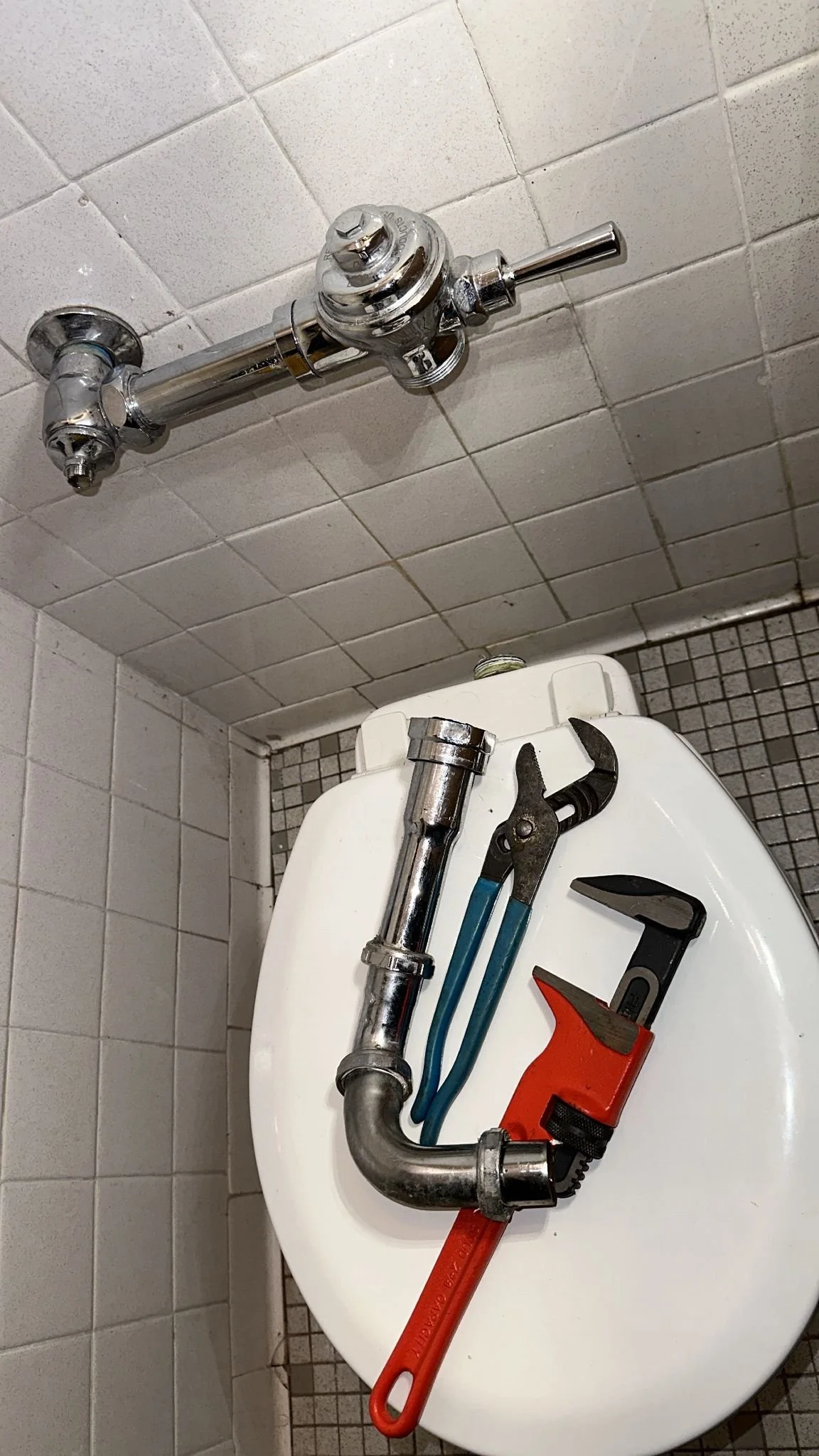 Tools including a pipe wrench, adjustable wrench, and pipe cutter placed on a toilet seat, with plumbing pipes and a water valve fixture on the tiled bathroom wall.