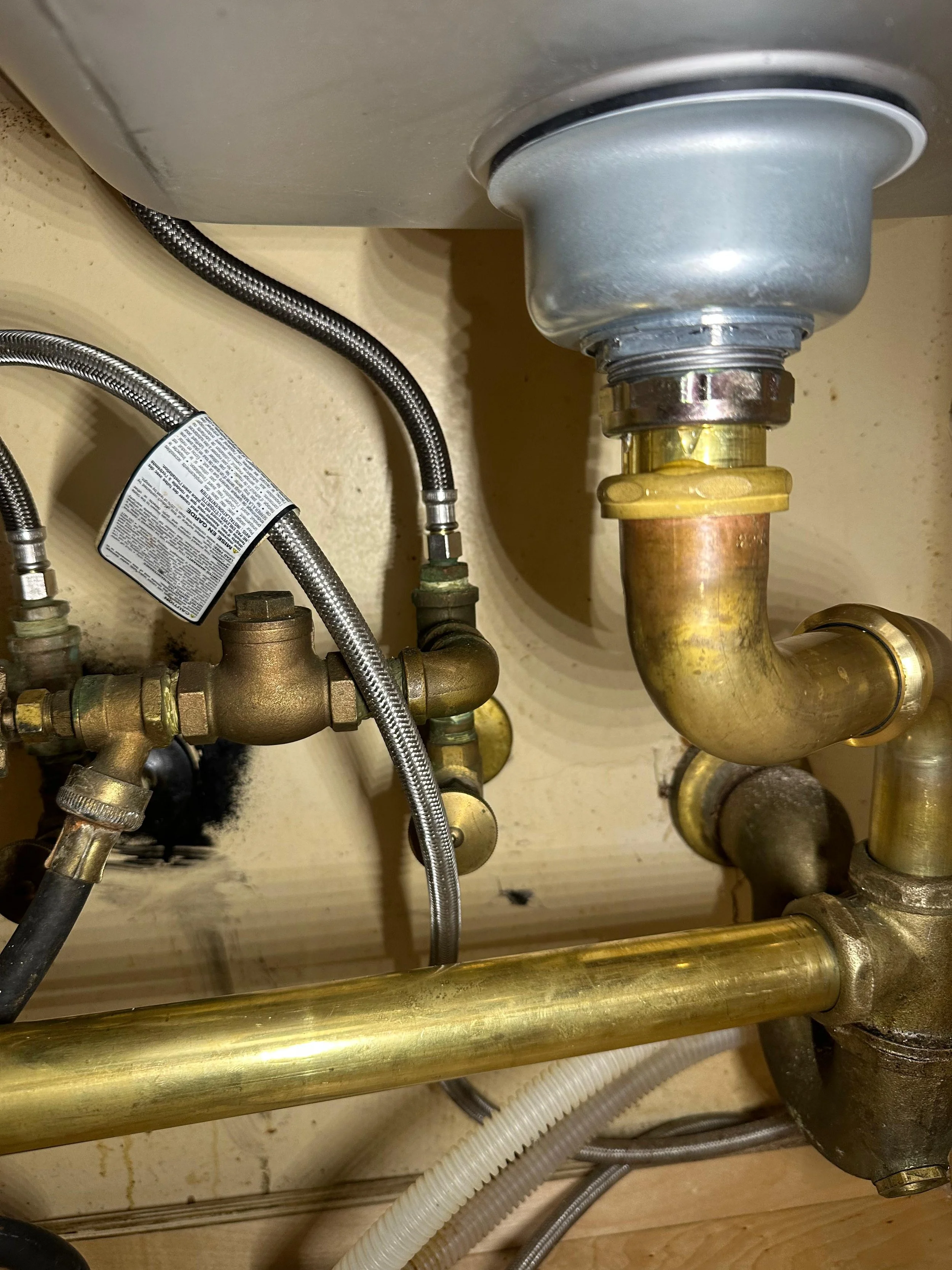 Underneath a kitchen sink, showing various pipes, including a brass pipe, a PVC pipe, and flexible hoses, with some black dirt or grime near the bottom.