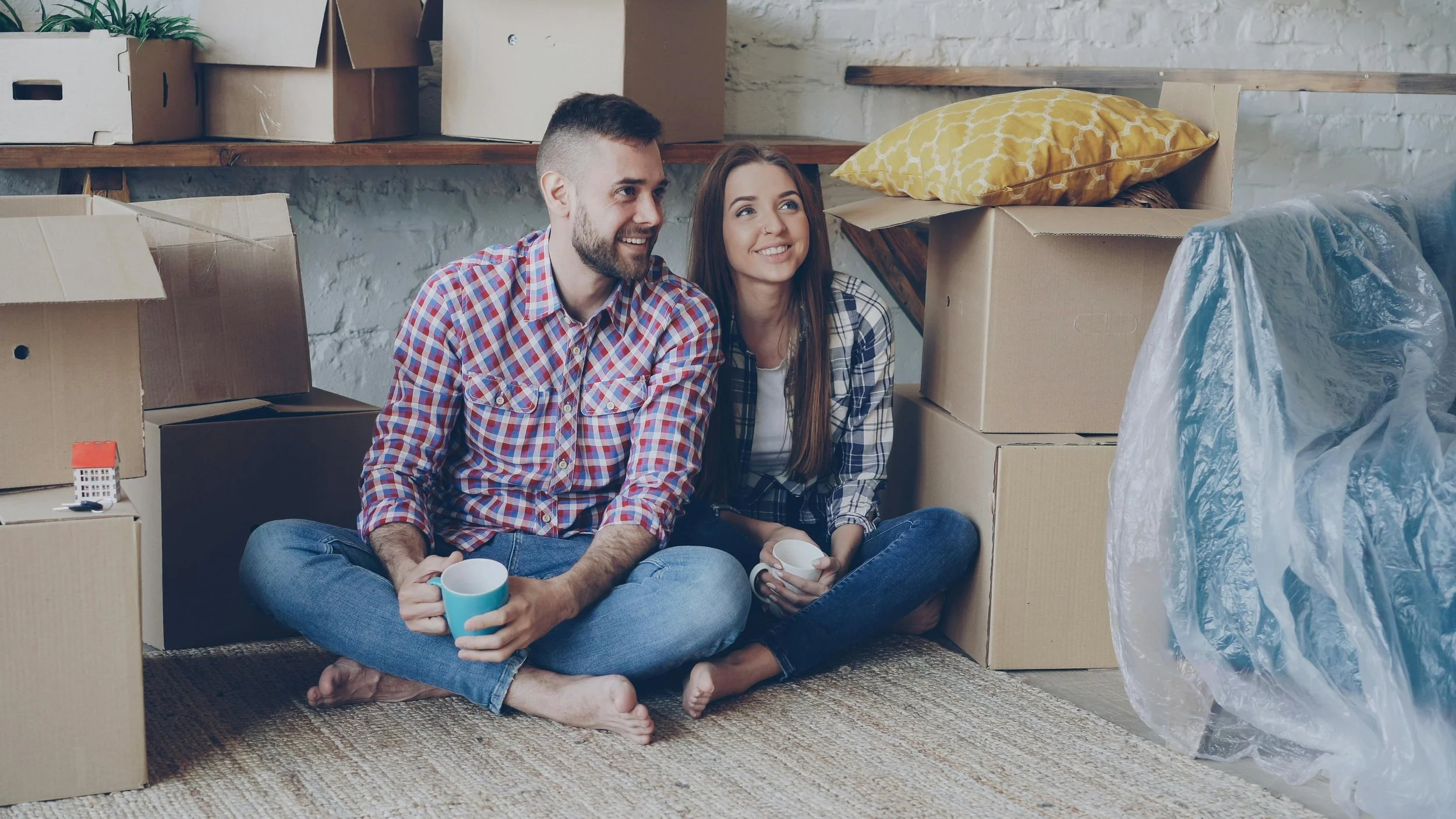 A couple who has just moved into a new rental apartment sit on the floor smiling, surrounded by boxes as they take a break from unpacking.