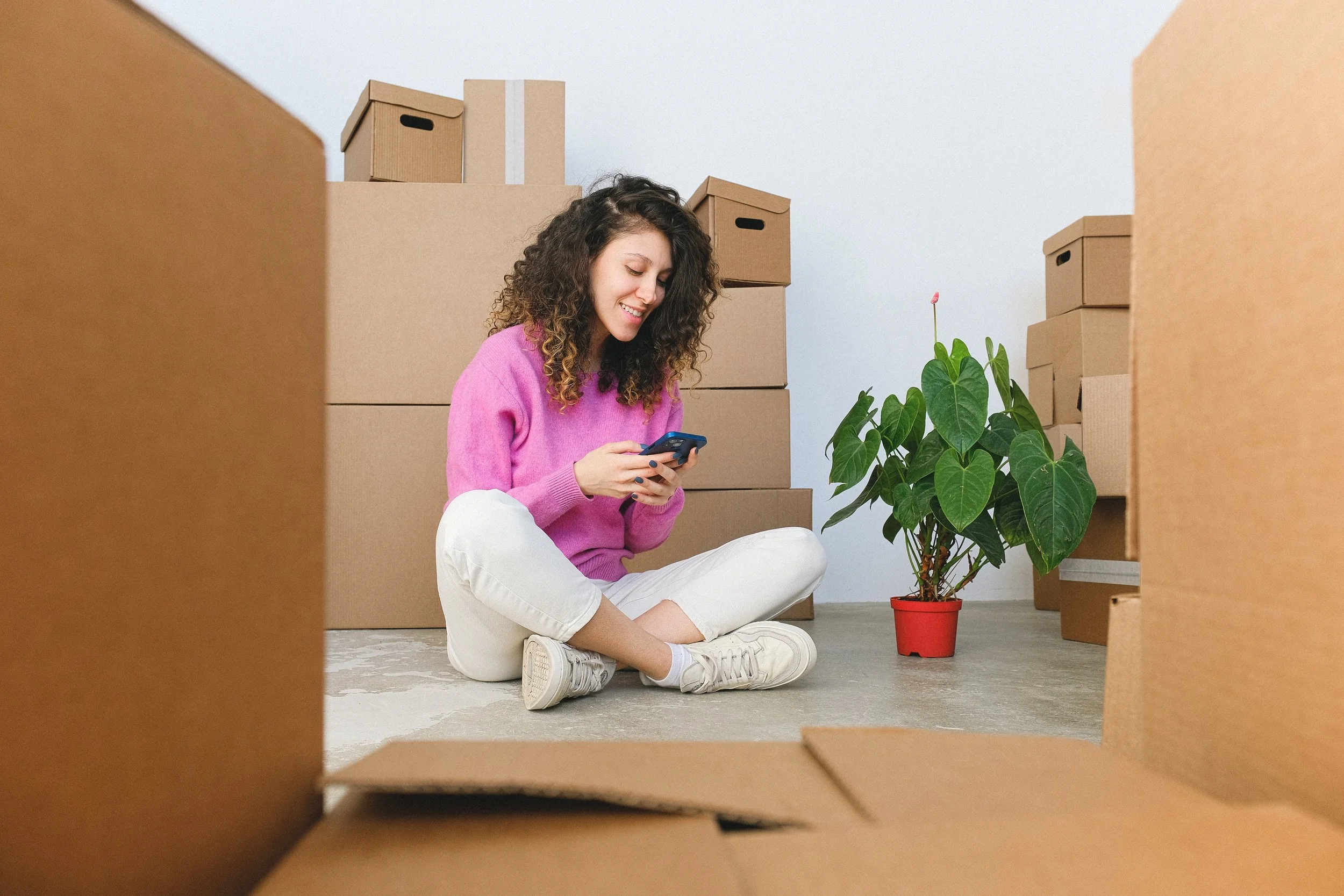 A young woman sits cross-legged on the floor next to a potted plant, framed by moving boxes, while looking, smiling, at a smartphone.