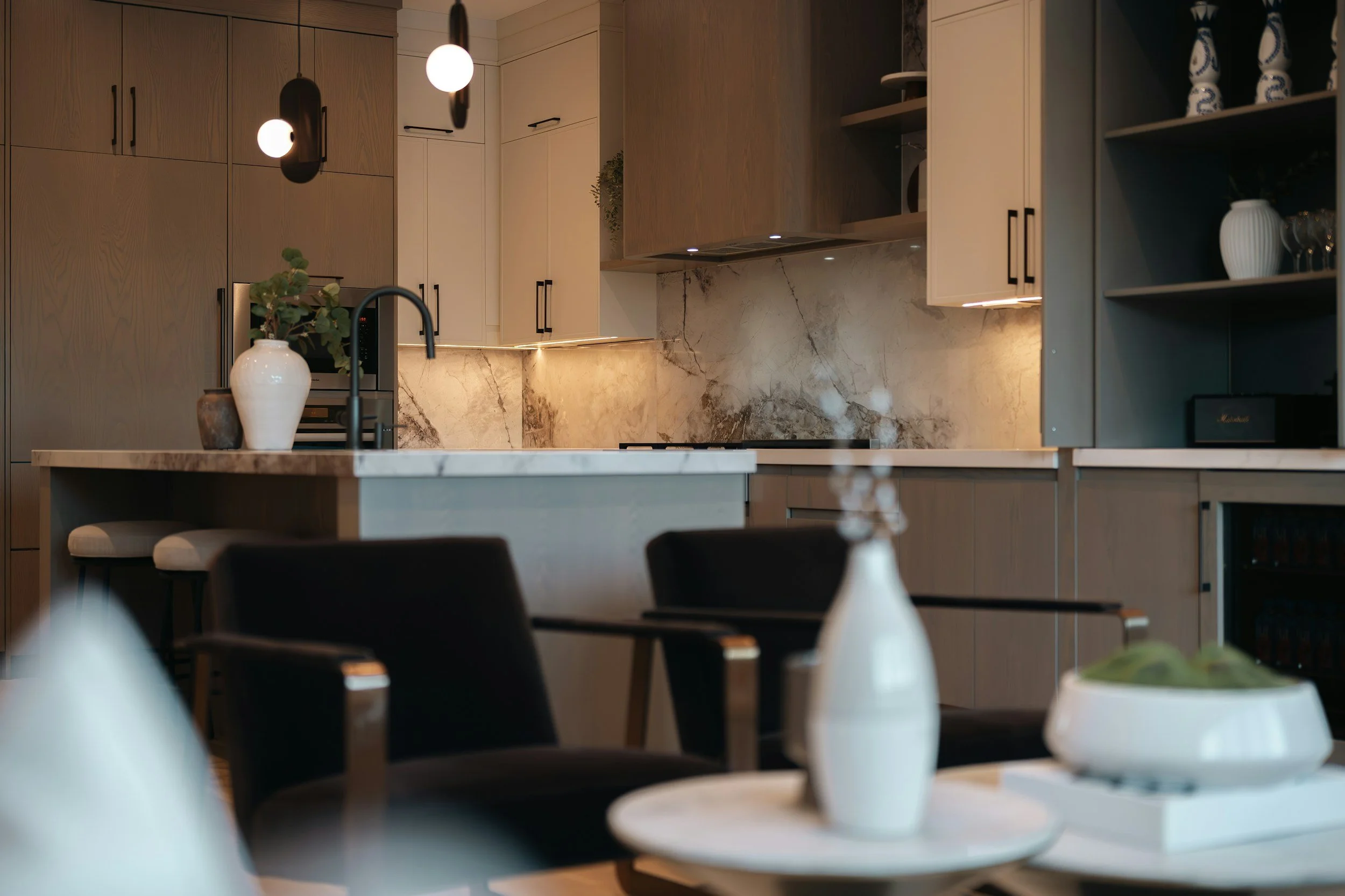 Modern kitchen with beige cabinets, marble backsplash, pendant lights, and decorative vases on the open shelves.
