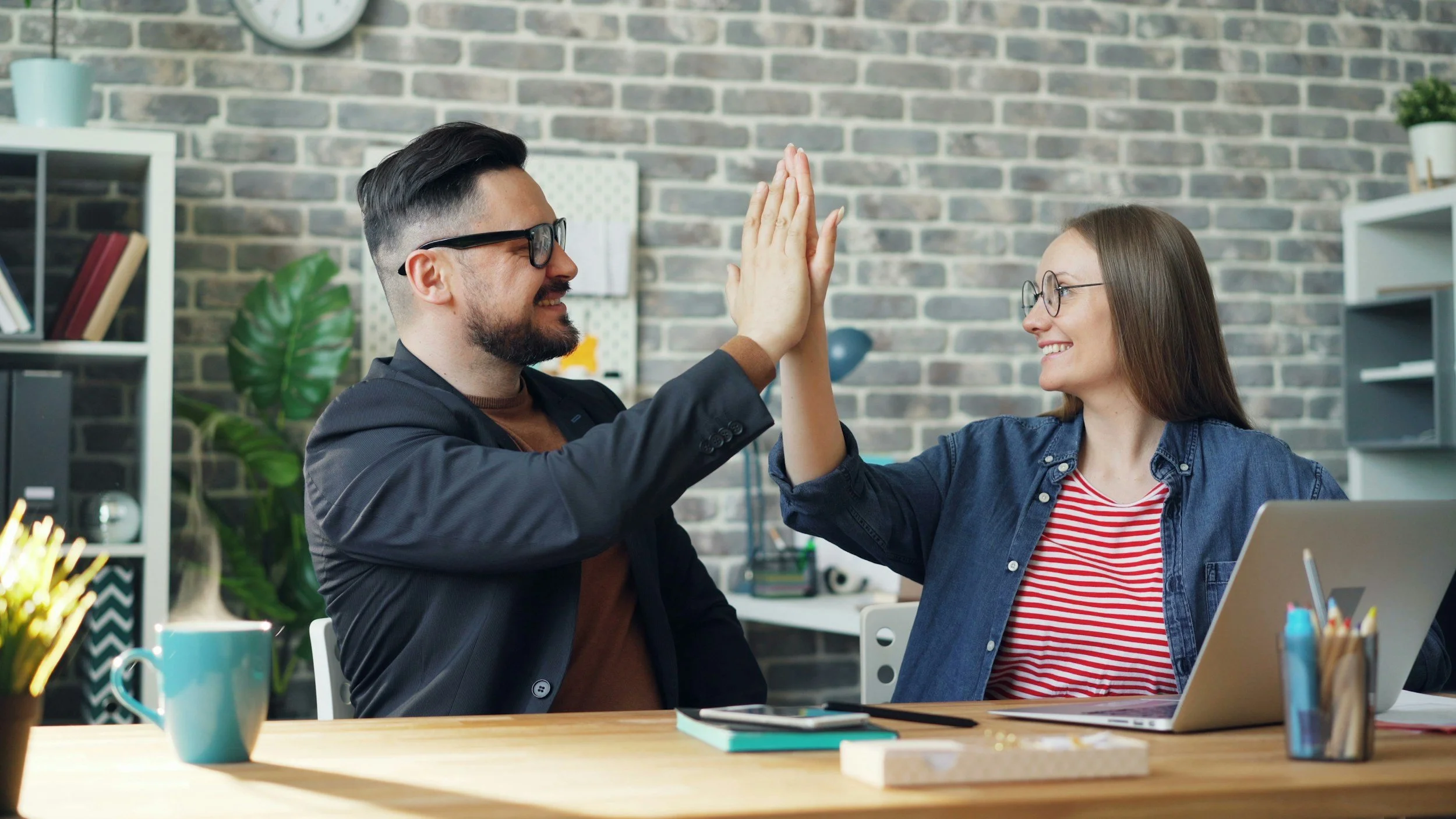 Two colleagues, a man and a woman, giving each other a high five and smiling in an office setting with a brick wall background.
