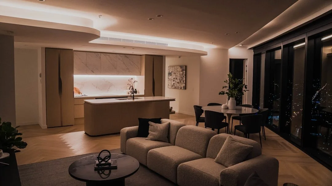Modern living room with beige sofa, small round coffee table, kitchen area with island, dining table with chairs, and large windows at night showing city lights.