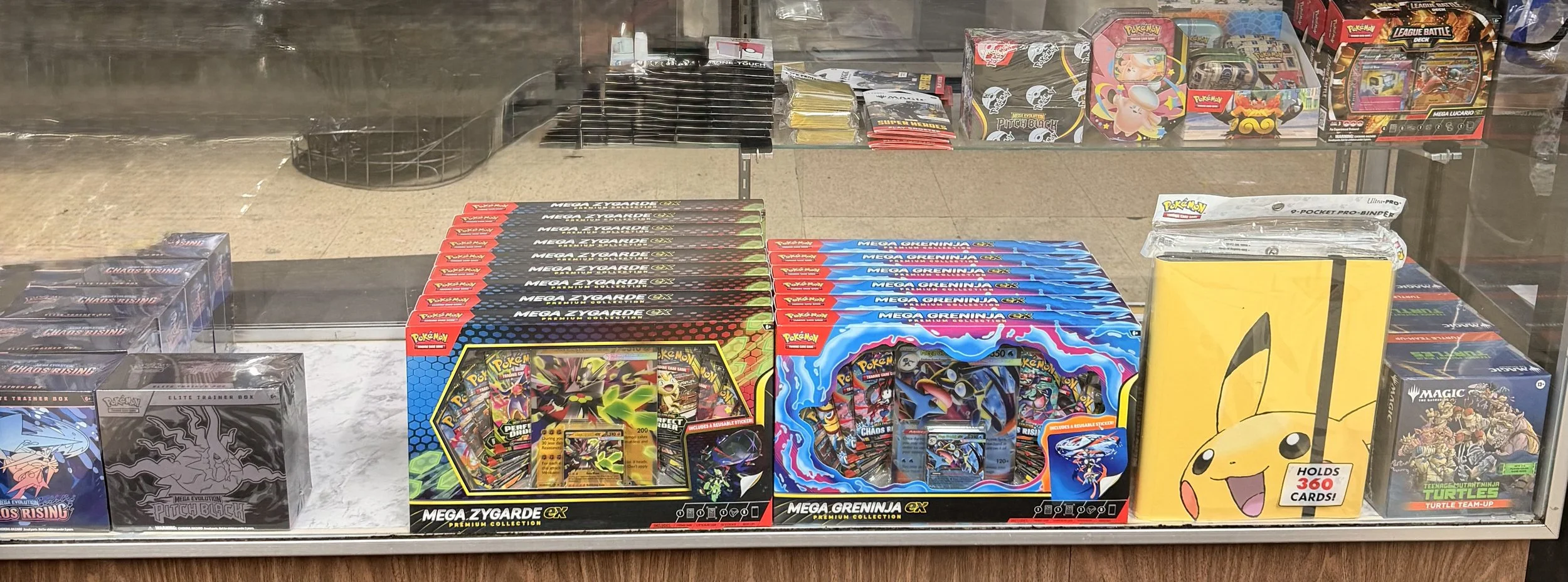 Display of Pokémon trading card booster boxes, including Mega Zygarde and Mega Greninja EX, with other Pokémon and Pokémon-themed products in the background.
