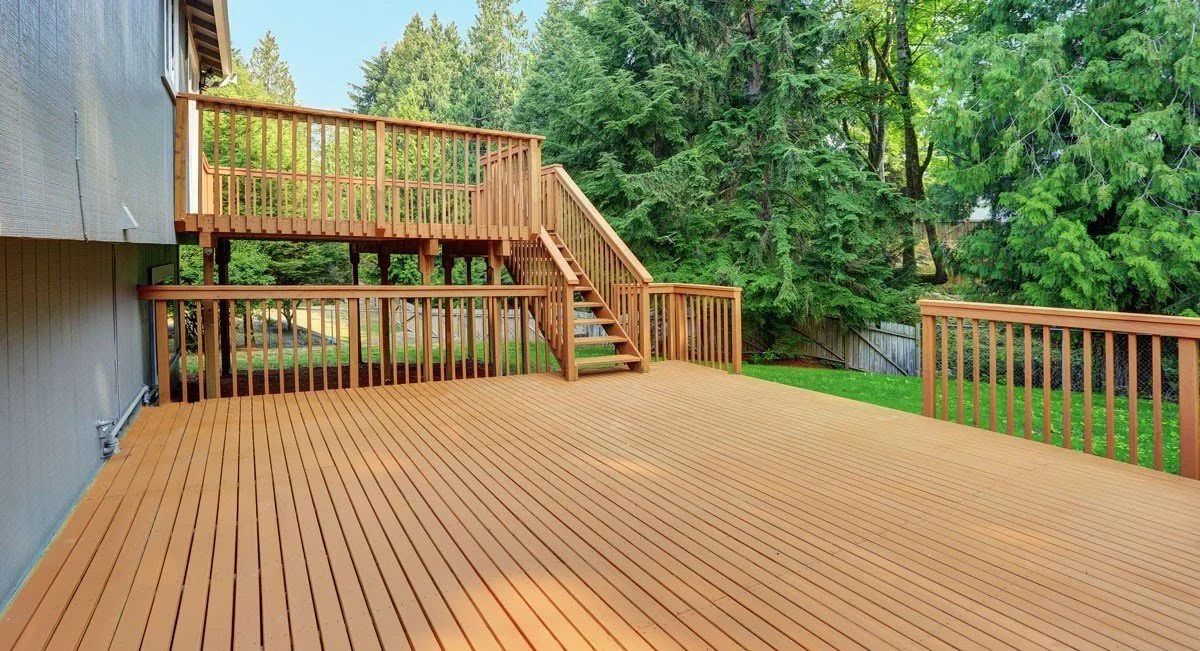 A large wooden deck attached to a house, with stairs leading up to a second-level deck, surrounded by green trees and a grass yard.