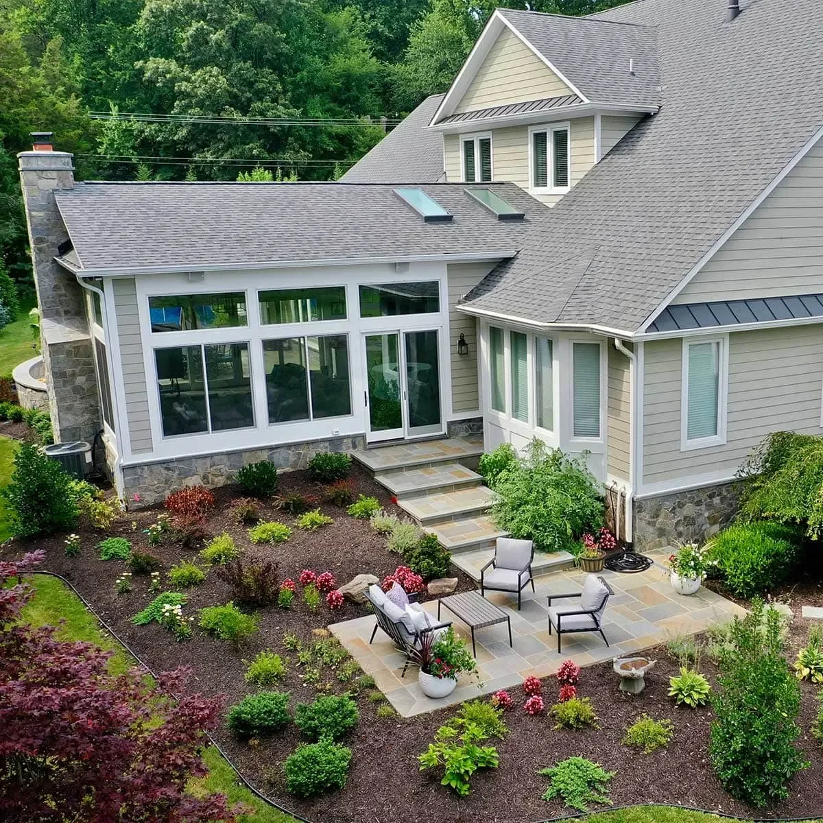 A house with a stone chimney, beige siding, and a gray shingle roof. It features large windows and skylights. There is a landscaped backyard with a patio, outdoor furniture, potted plants, and a garden with various shrubs and flowers.