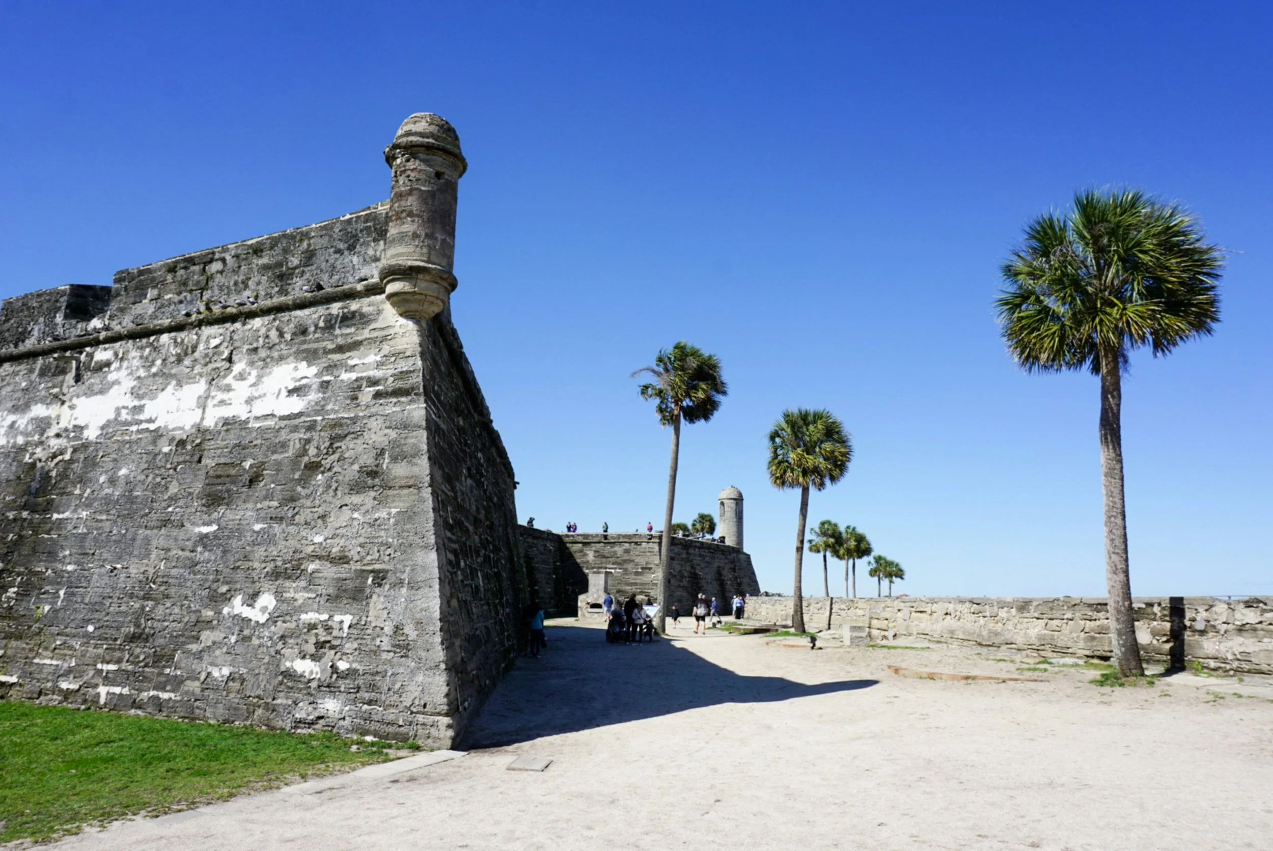 Historical stone fortress wall with a rounded turret and palm trees under a clear blue sky, with people walking and sitting nearby.